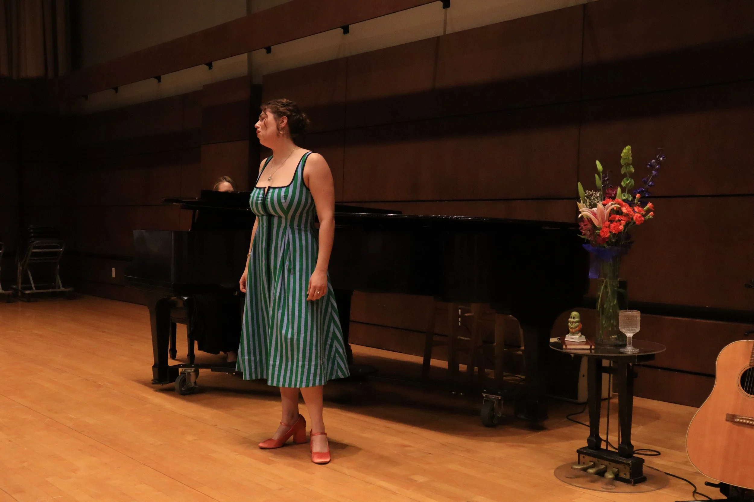 A woman in a green and white striped dress and high heels stands on a stage with a piano and a bouquet of flowers nearby.