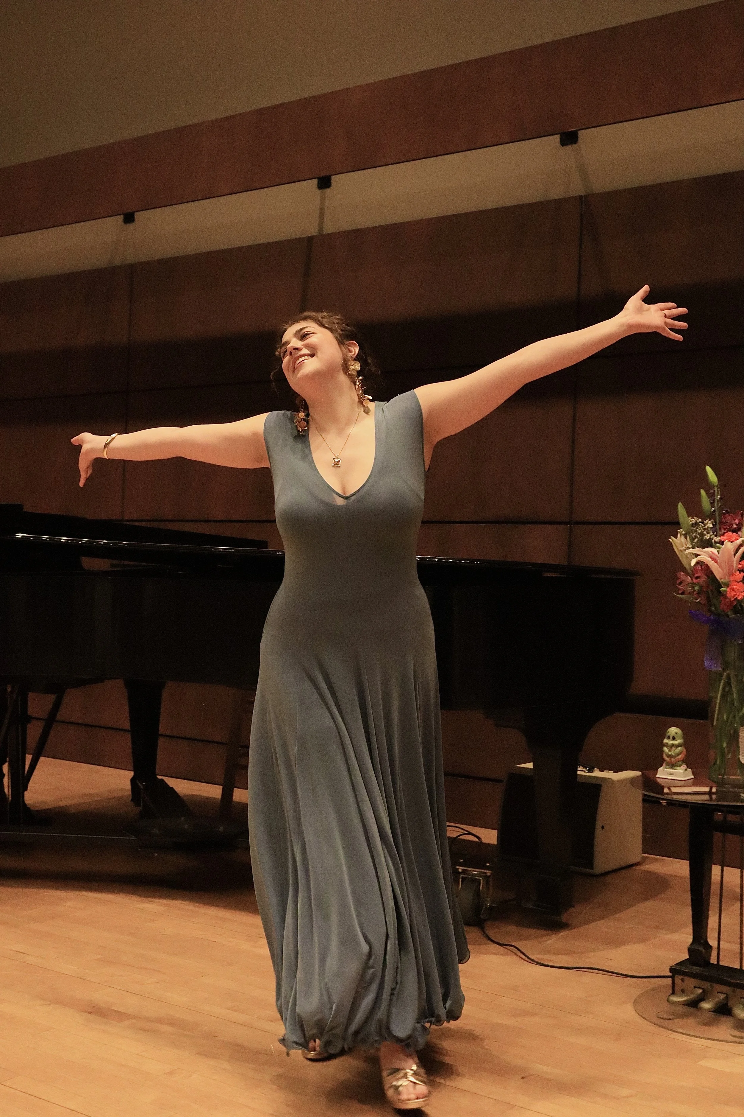 A woman in a long gray dress dancing with arms outstretched in a music room with a grand piano, flowers, and decorative objects.