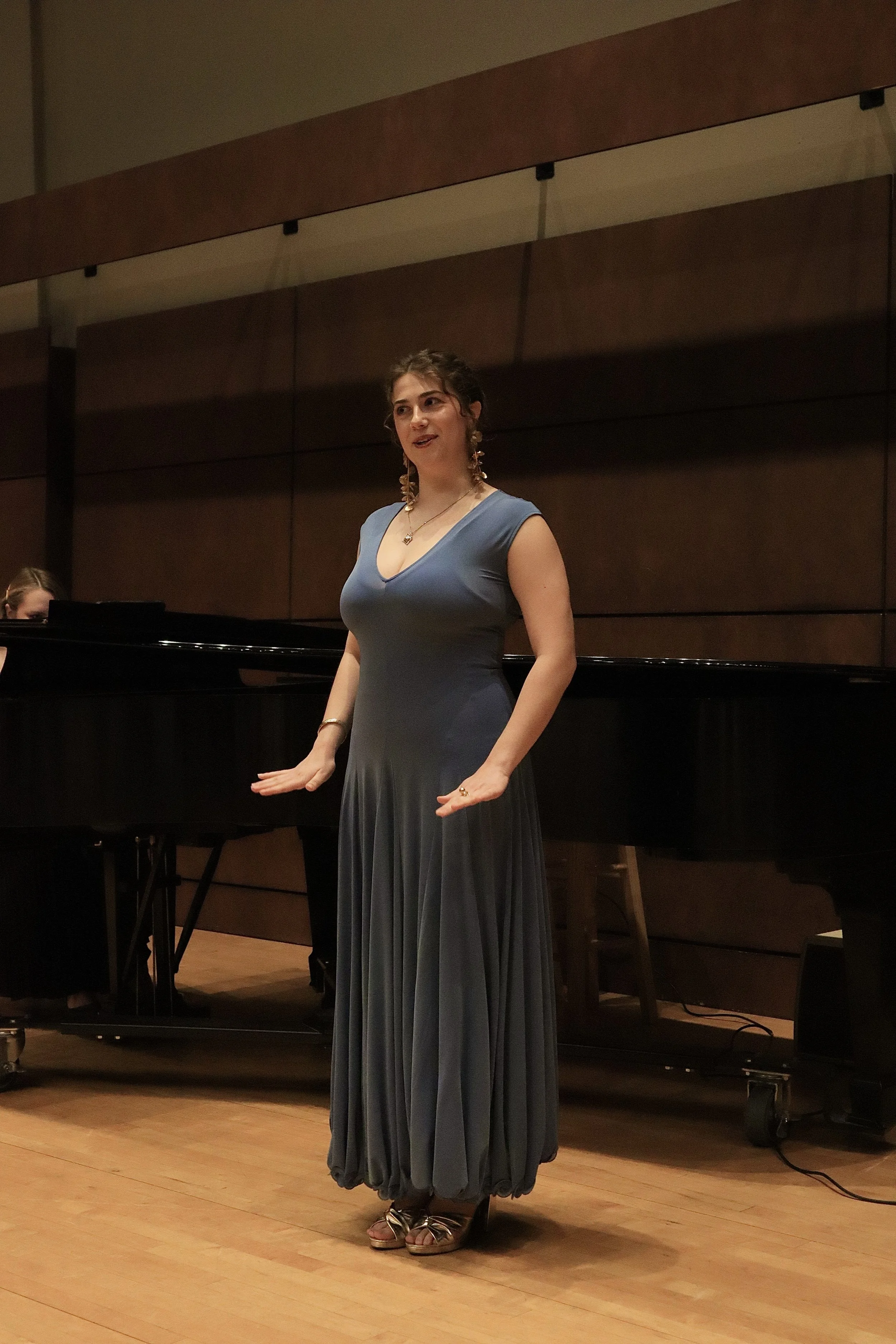 A woman in a long, flowing blue dress and gold heeled sandals standing on a wooden stage, possibly singing or addressing an audience, with a grand piano and another musician partially visible in the background.