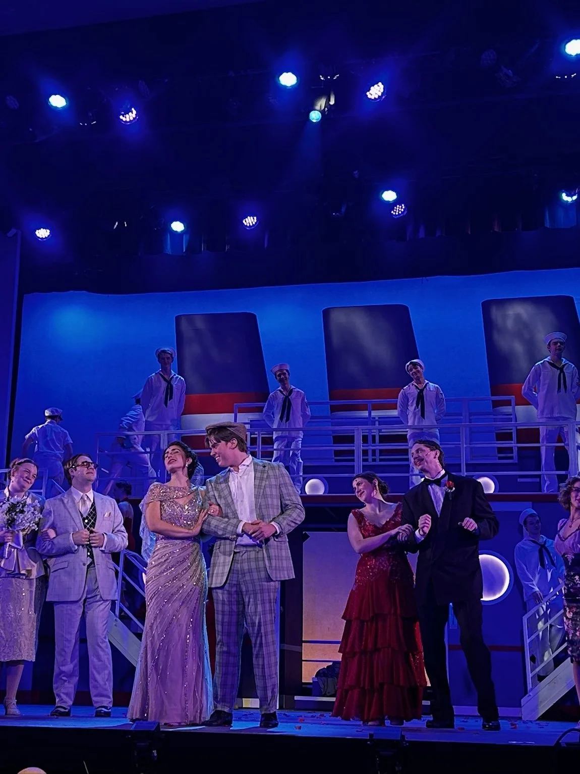 Stage scene from a musical or play with performers in vintage costumes, including sailors and formal wear, under blue lighting.