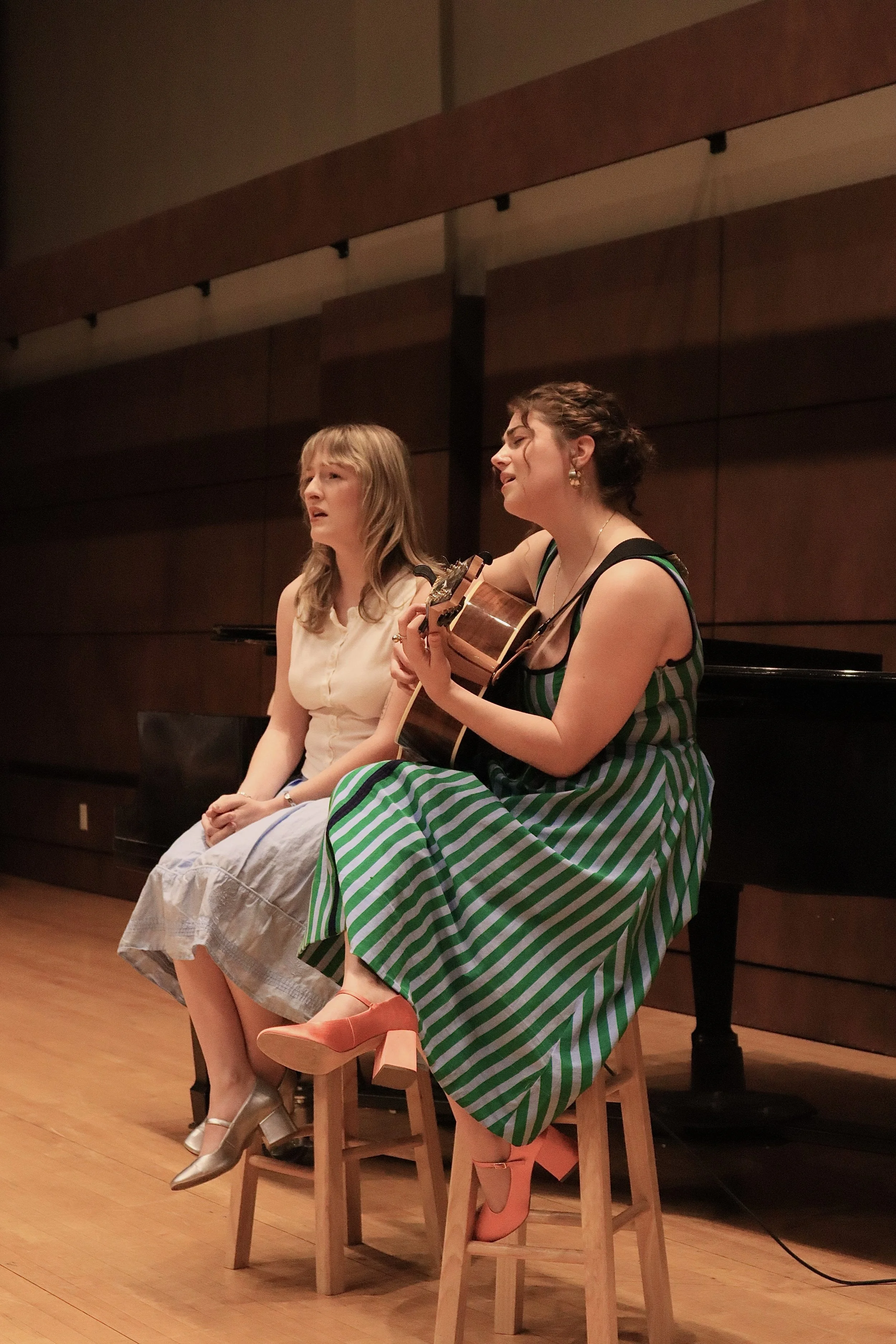 Two women sitting on stools, one playing a guitar and singing, the other singing. They are on a stage with wooden floors and a dark wooden wall background.