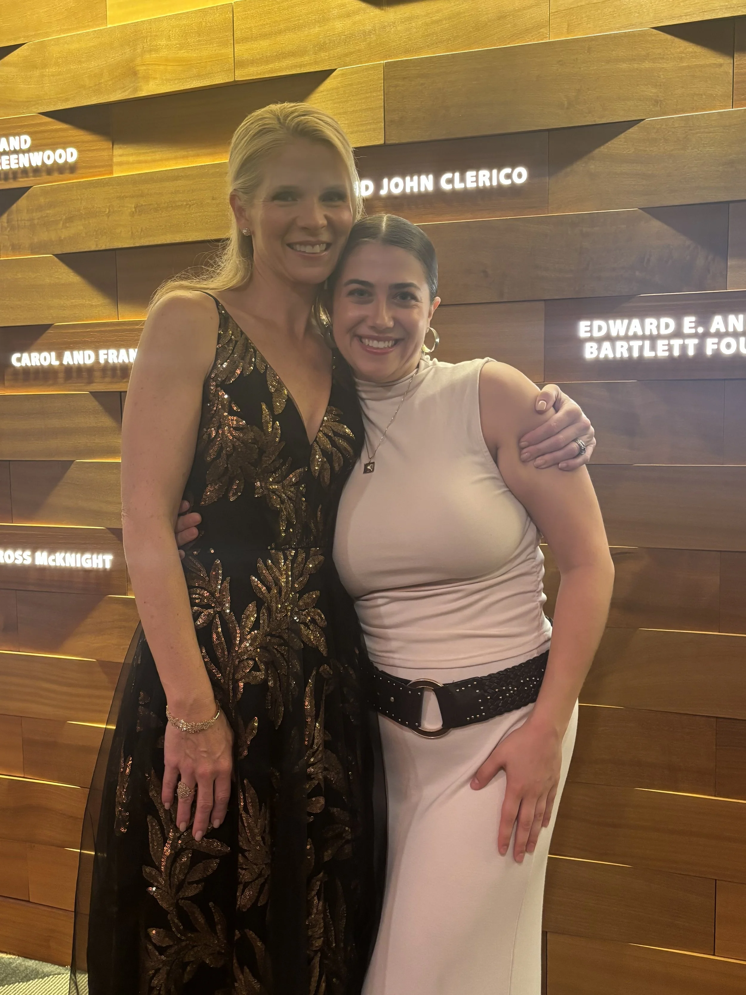 Two women smiling and embracing in front of a wooden wall with illuminated text plaques.