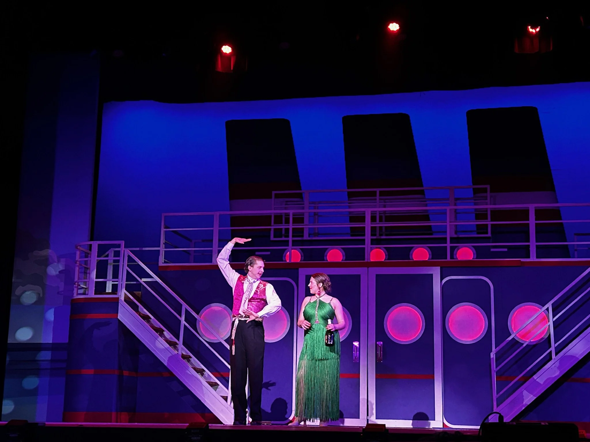 Two actors performing on a stage with a colorful, abstract background and circular light design. One actor is wearing a pink vest and black trousers, the other is in a green dress. Both are engaged in conversation, with the man gesturing upward.