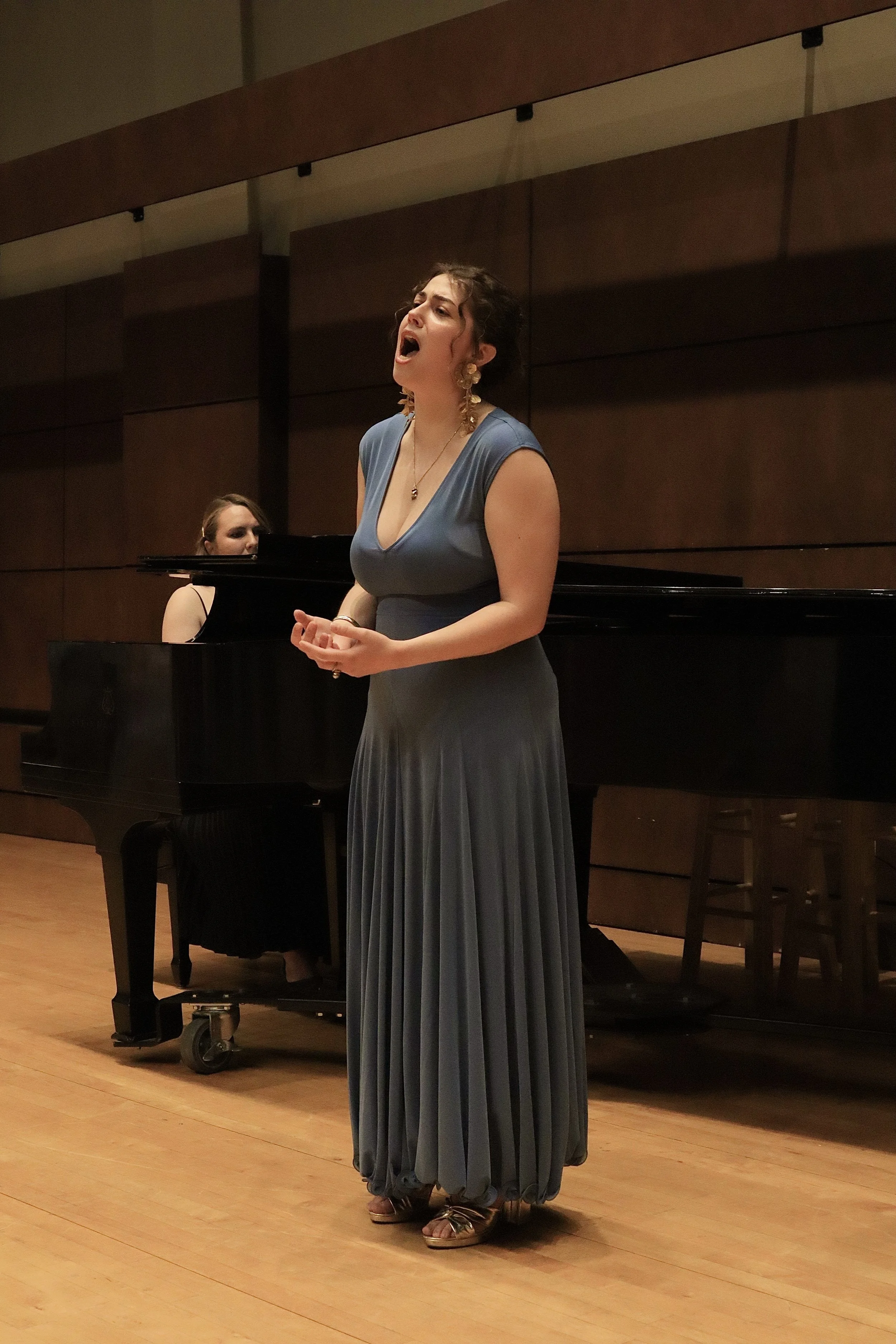 A woman singing with her eyes closed, wearing a long blue dress and gold jewelry, standing near a grand piano in a performance hall.