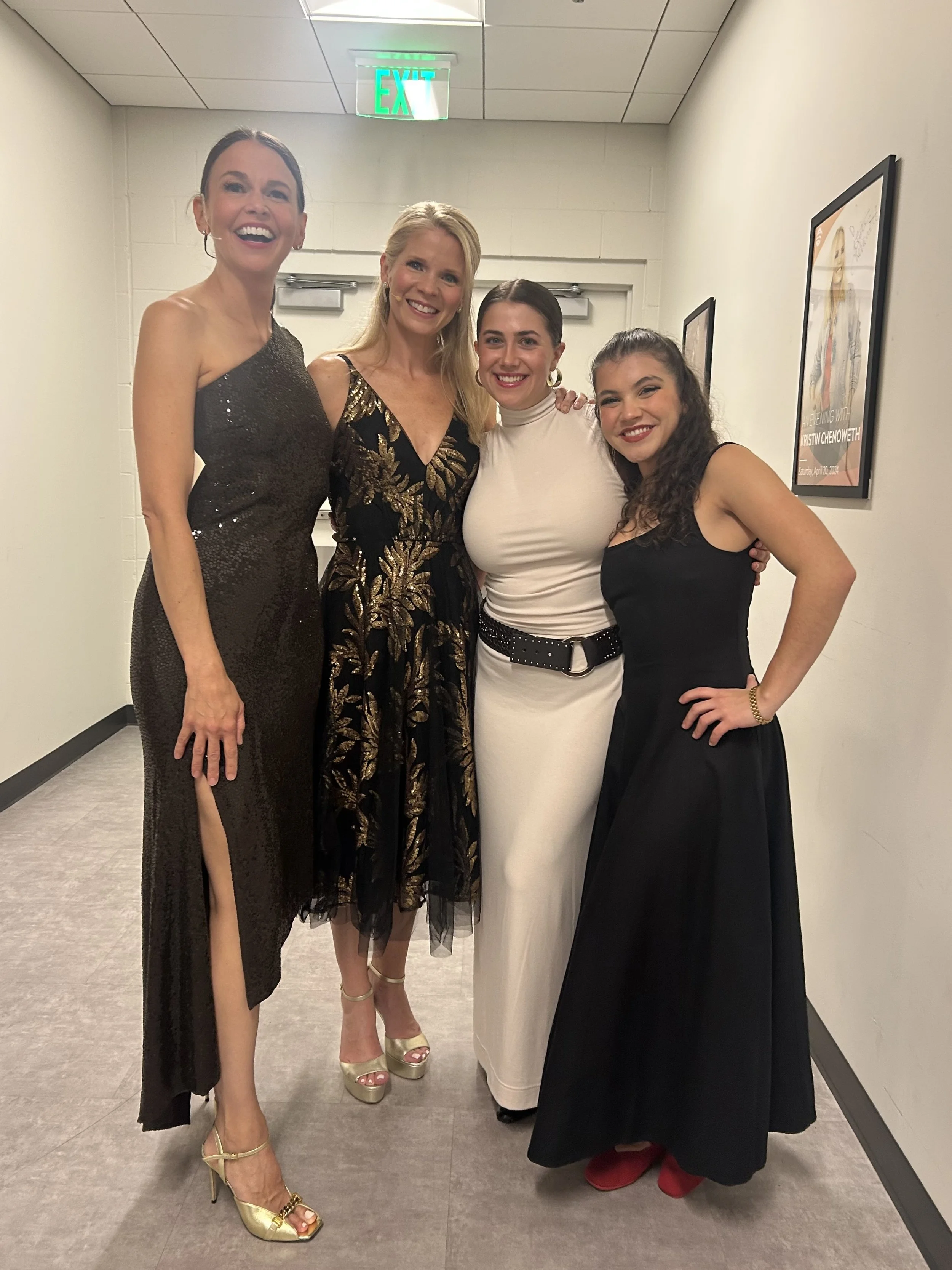 Four women dressed in elegant evening attire standing close together in a hallway, smiling for the camera. The woman on the far left is wearing a black sequined dress with a high slit and gold high-heeled shoes. The second woman is wearing a black an