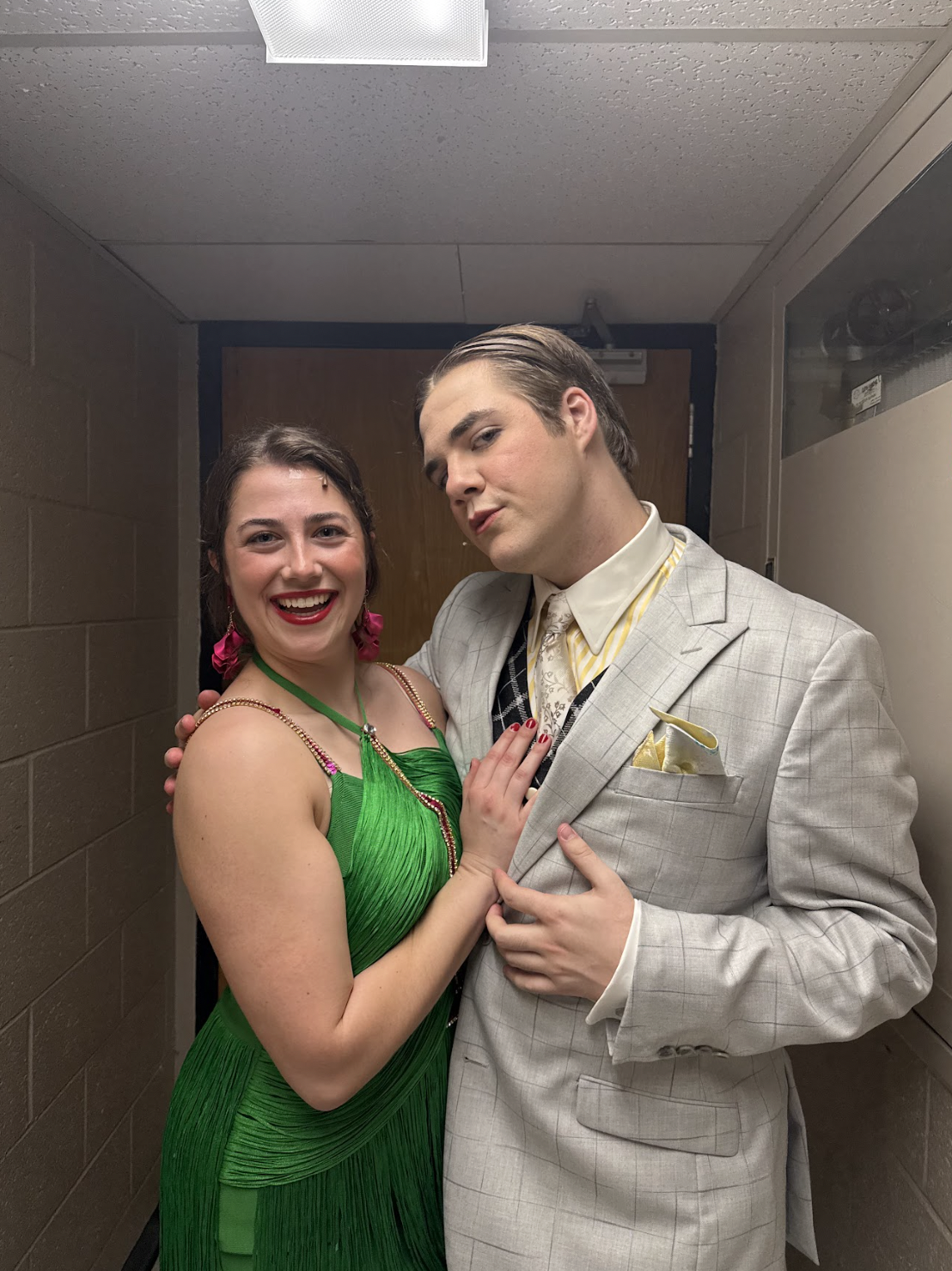 A woman in a green dress and a man in a light patterned suit pose together, smiling and looking at the camera, in a narrow hallway with a wooden door behind them.