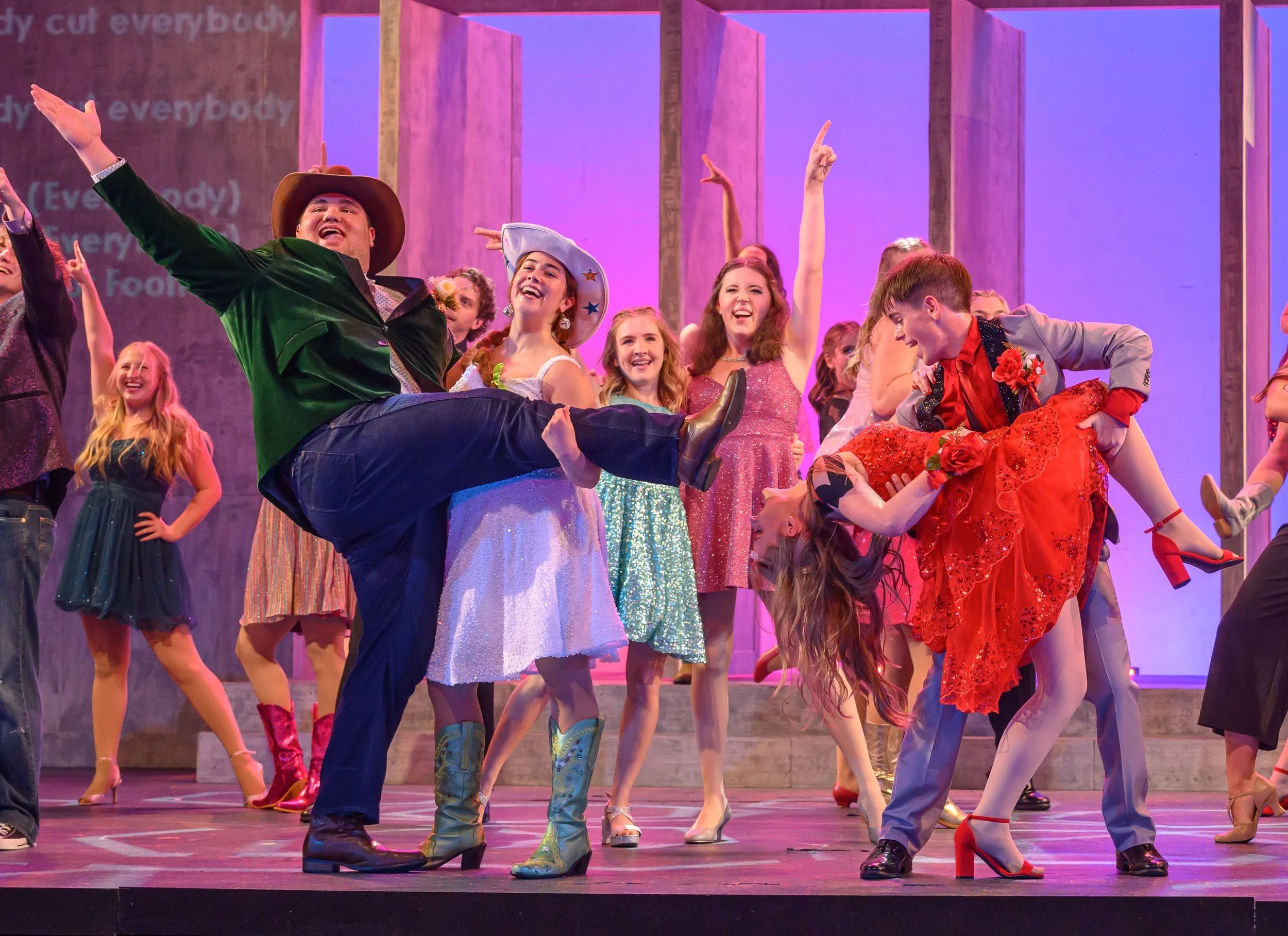 A group of performers on stage, dressed in colorful costumes, celebrating and dancing during a theatrical production. The background features purple lighting and wooden set pieces.