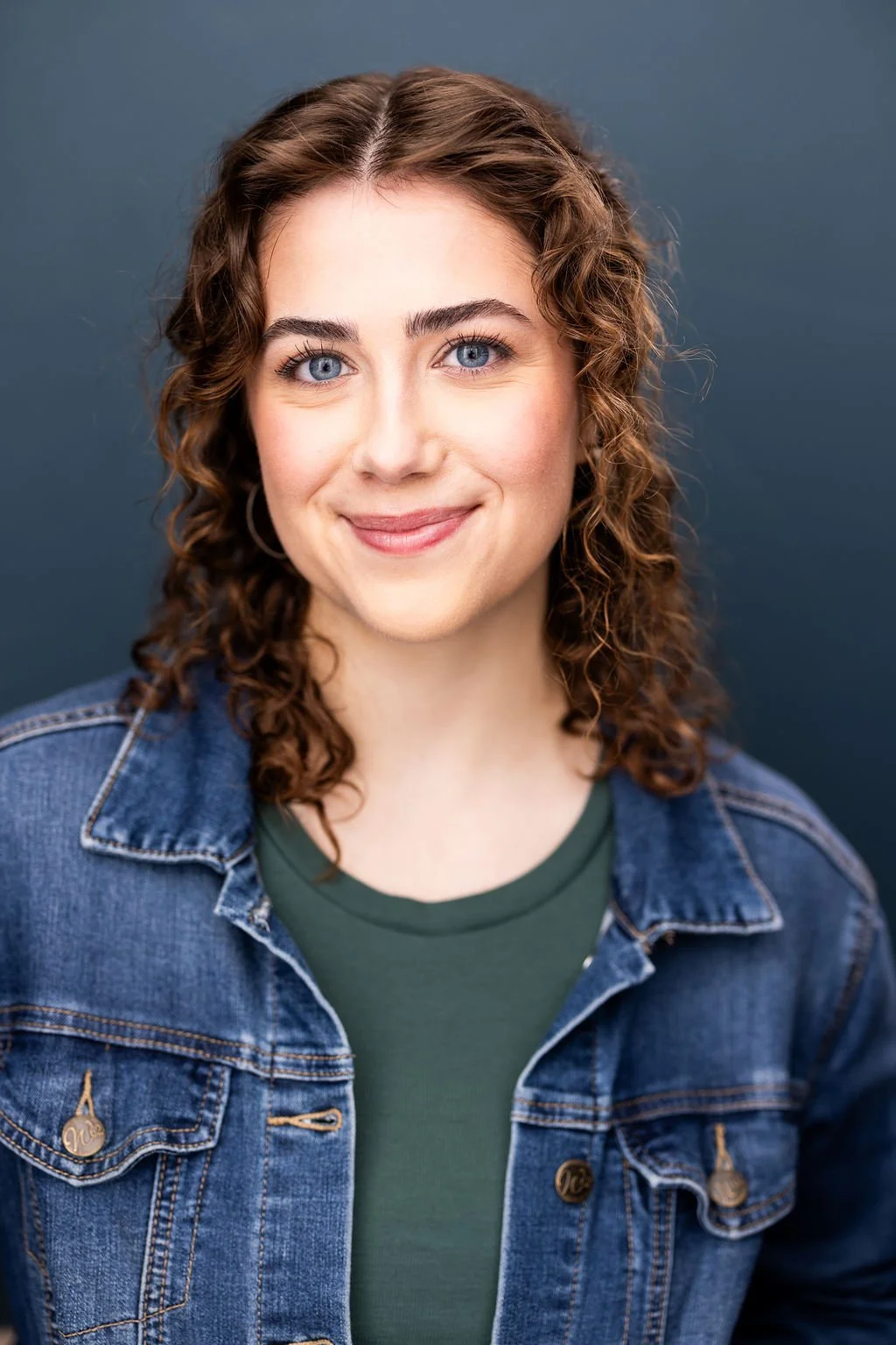 A young woman with curly brown hair, blue eyes, and a warm smile, wearing a denim jacket over a green shirt, standing against a dark gray background.