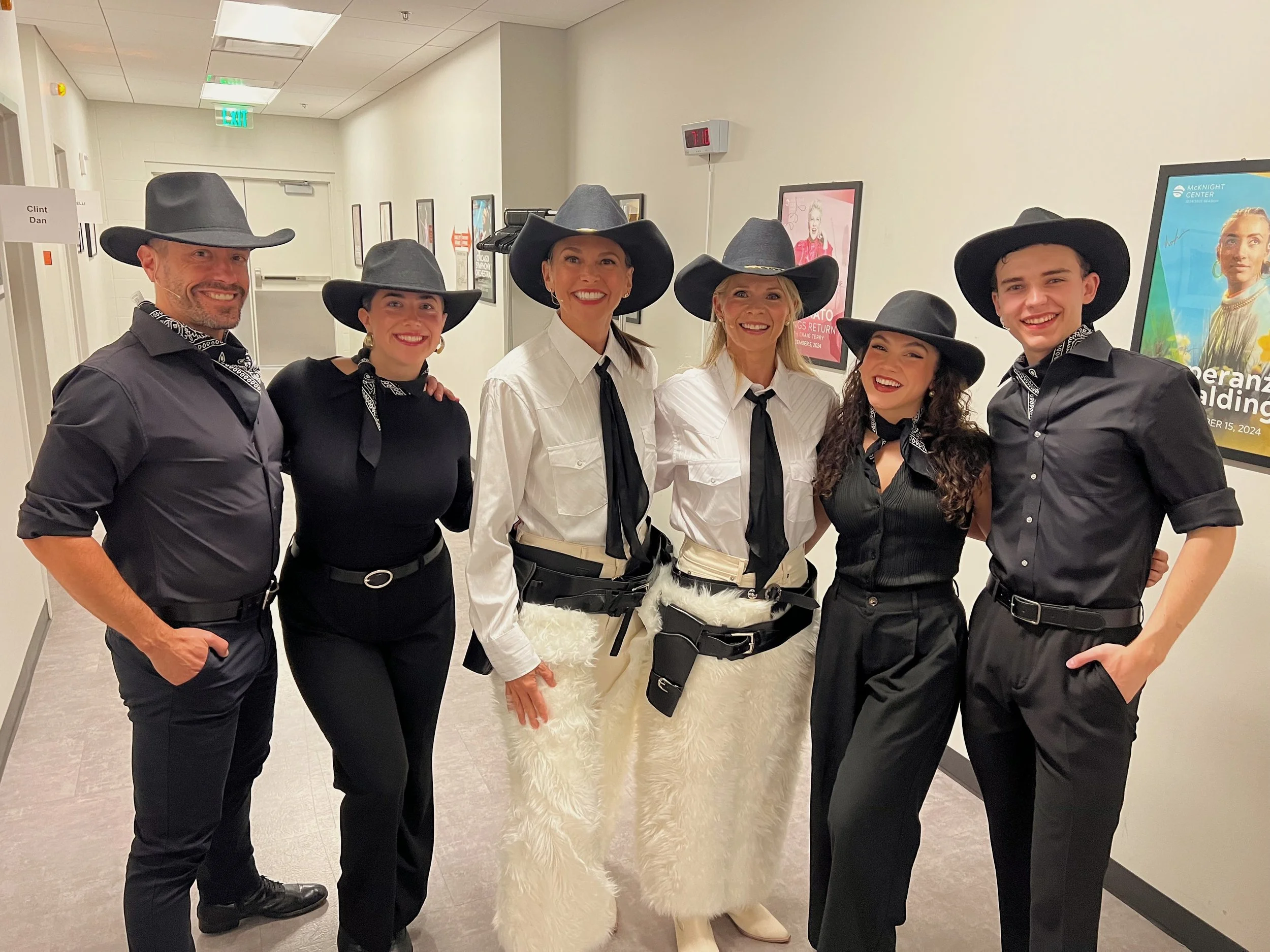 Group of six people dressed in cowboy and cowgirl costumes, wearing black hats and bandanas, standing in a hallway with framed posters on the wall.