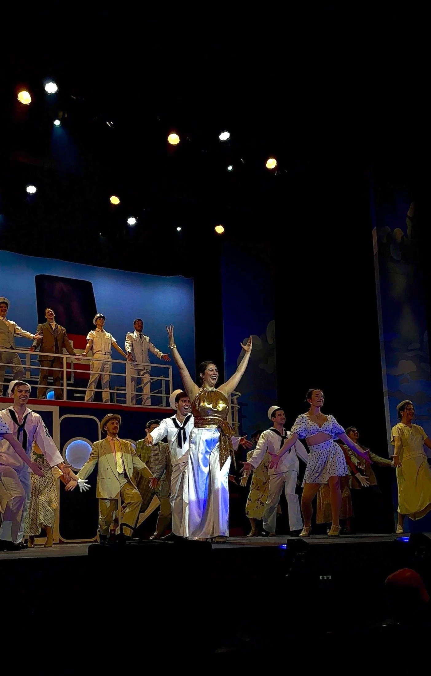 A theatrical stage performance with a group of actors dressed in vintage costumes, some in sailor outfits and others in polka-dot dresses, celebrating with smiles and dance. A woman in the center, wearing a gold top and white pants, raises her arms i