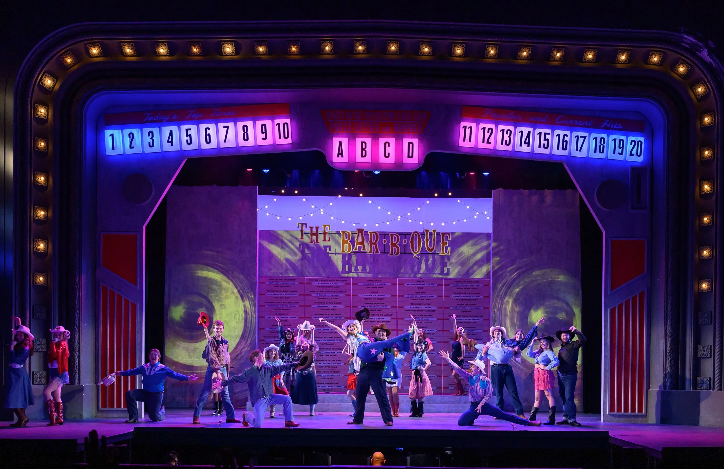 A stage performance of a musical show with actors dressed as cowboys and cowgirls, with a colorful set design and signs indicating game options above, under purple and blue stage lighting.