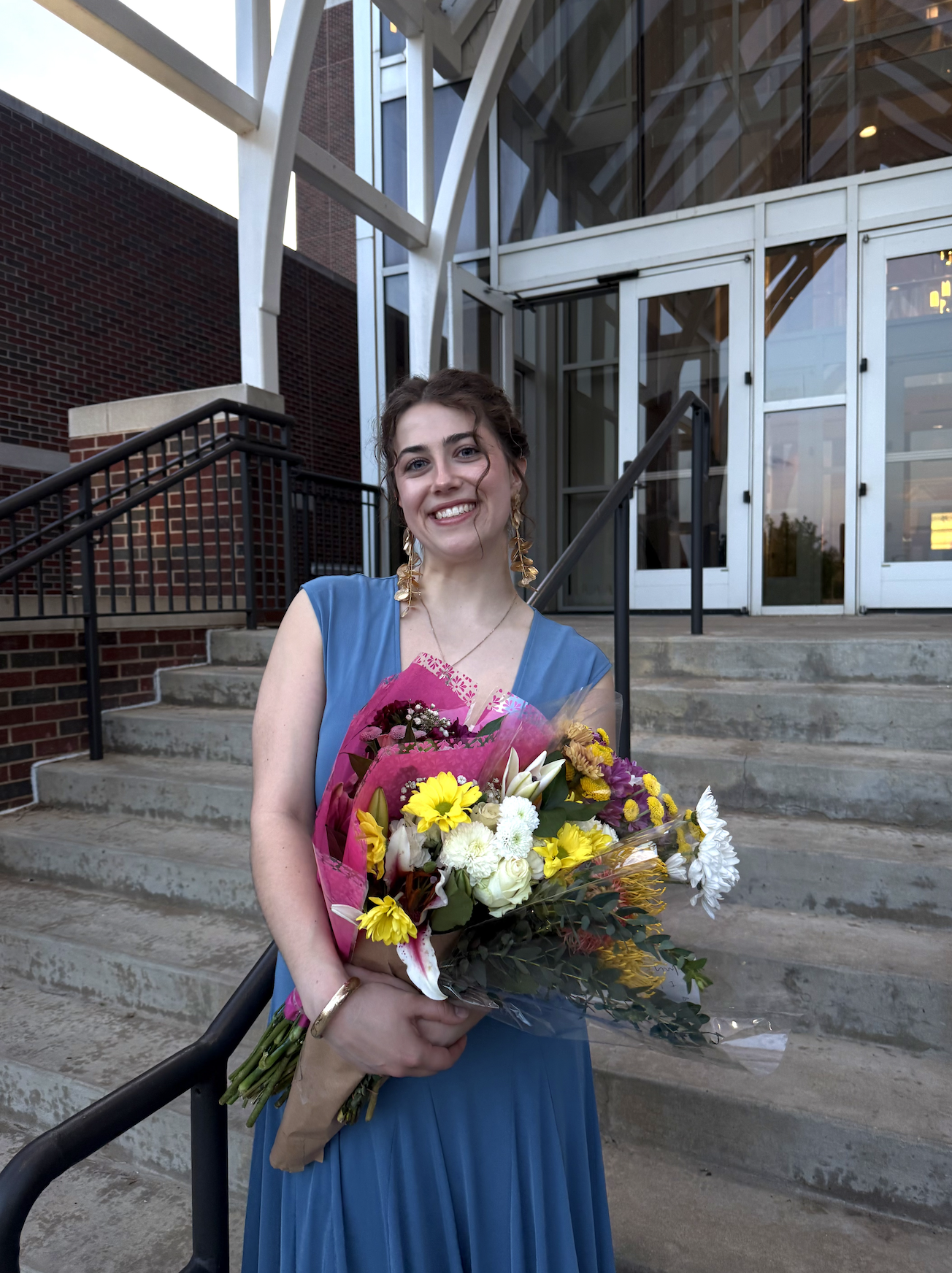A woman in a blue dress holding a bouquet of colorful flowers standing on concrete steps outside a modern building with glass and metal architecture.