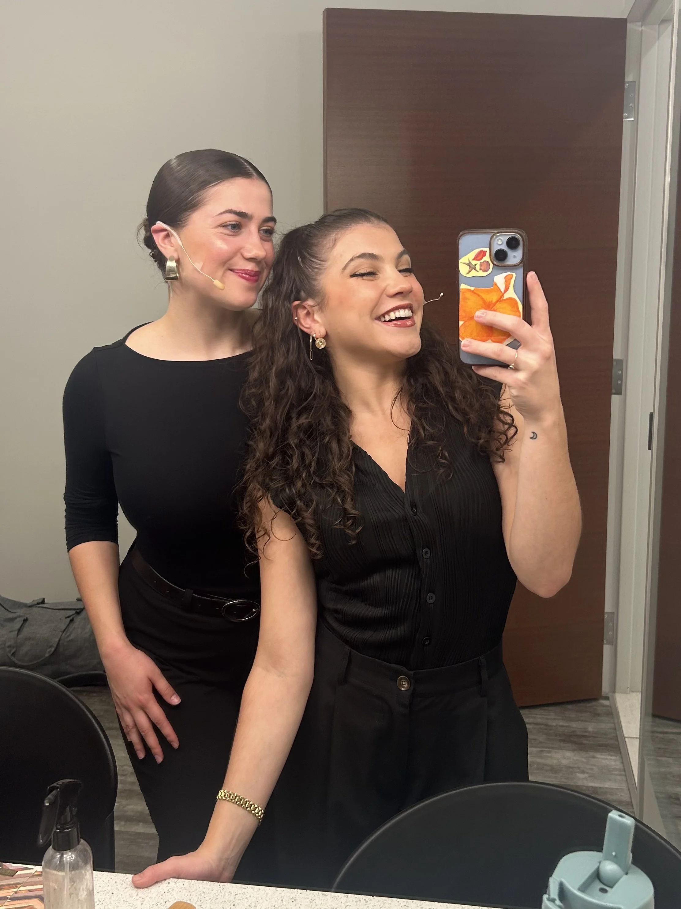 Two women taking a selfie in a dressing room bathroom. One woman has dark curly hair and is smiling with her eyes closed, wearing a black top. The other woman has dark straight hair, wearing a headset microphone, earrings, a black top, and a gold bra