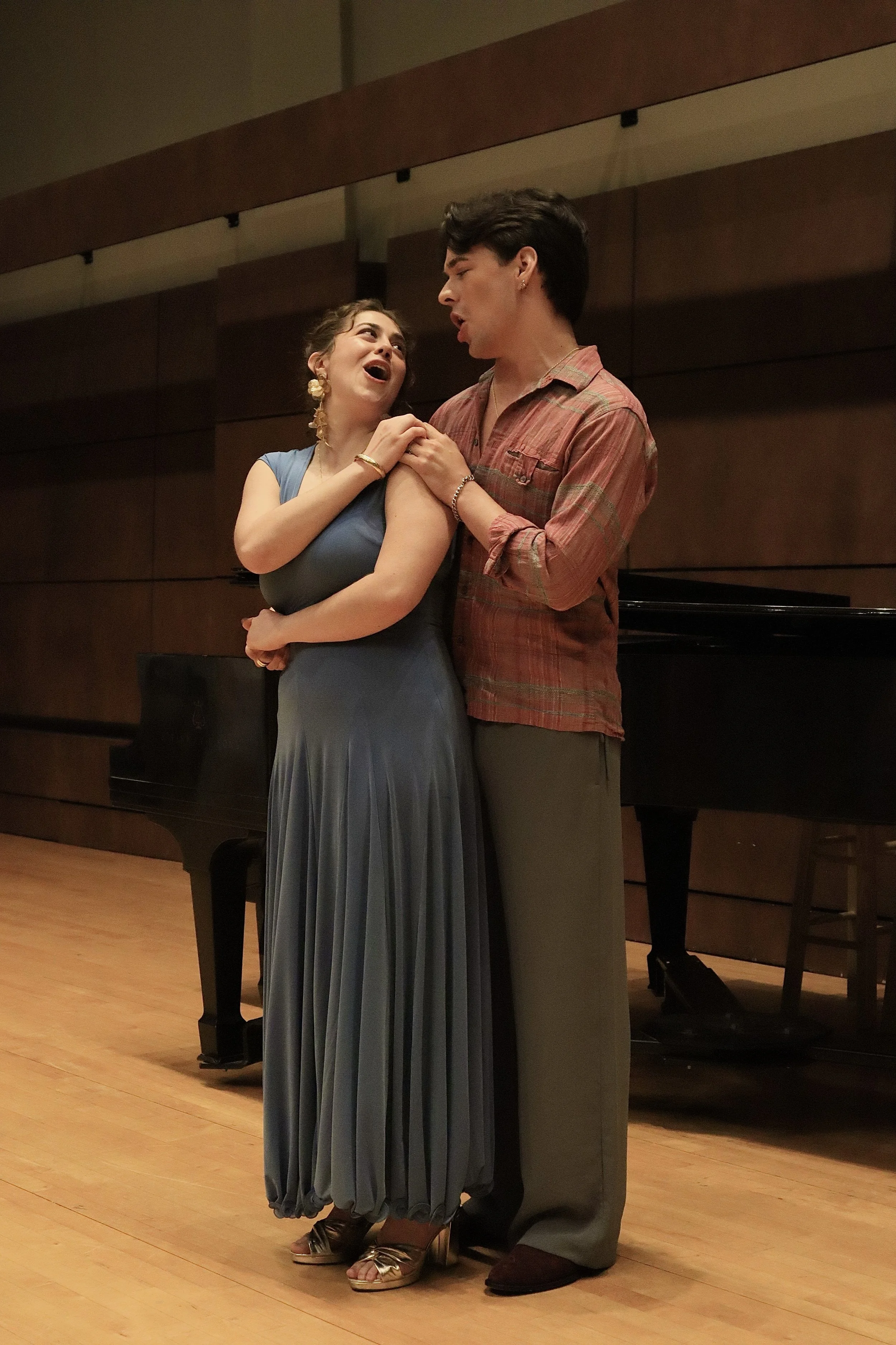 A man and woman are dancing together in a room with wooden floors and a grand piano in the background. The woman is wearing a blue dress and gold heels, and the man is wearing a red plaid shirt and gray pants.