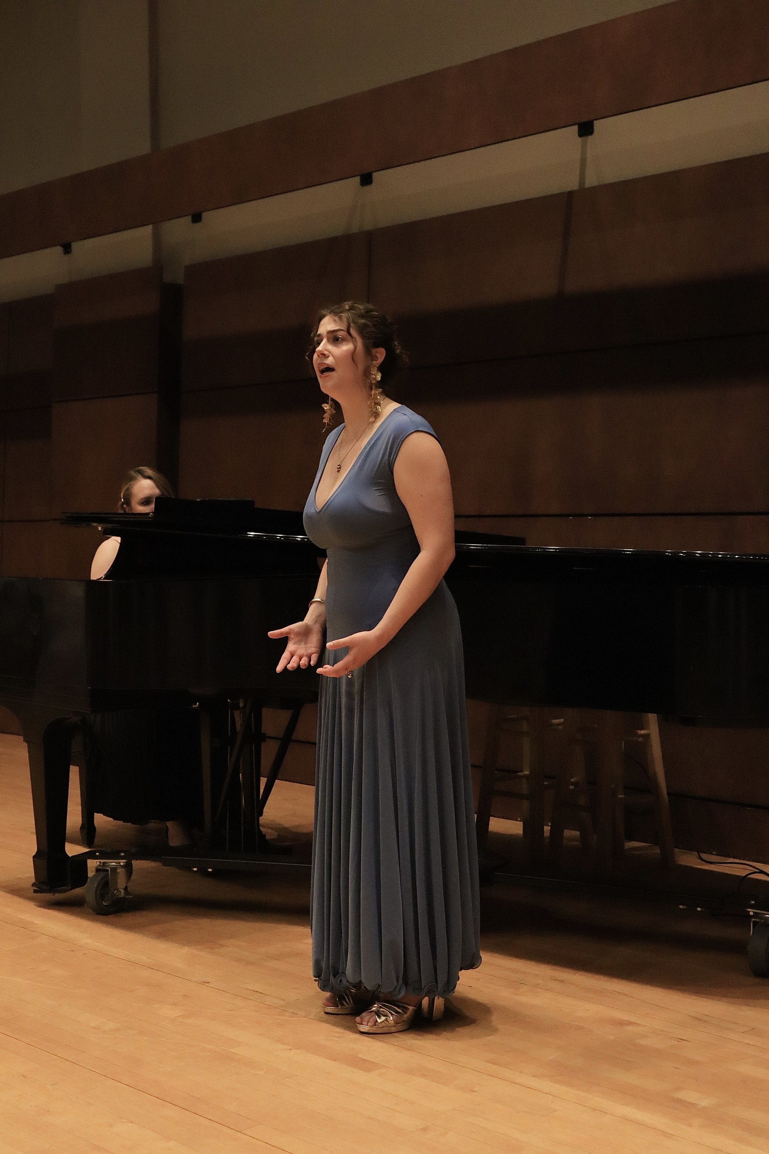 A woman in a long, elegant blue dress is performing in front of a piano, with a pianist seated behind her. The woman has dark hair pulled back, is wearing earrings, and appears to be singing or speaking passionately with her hands gesturing. The sett