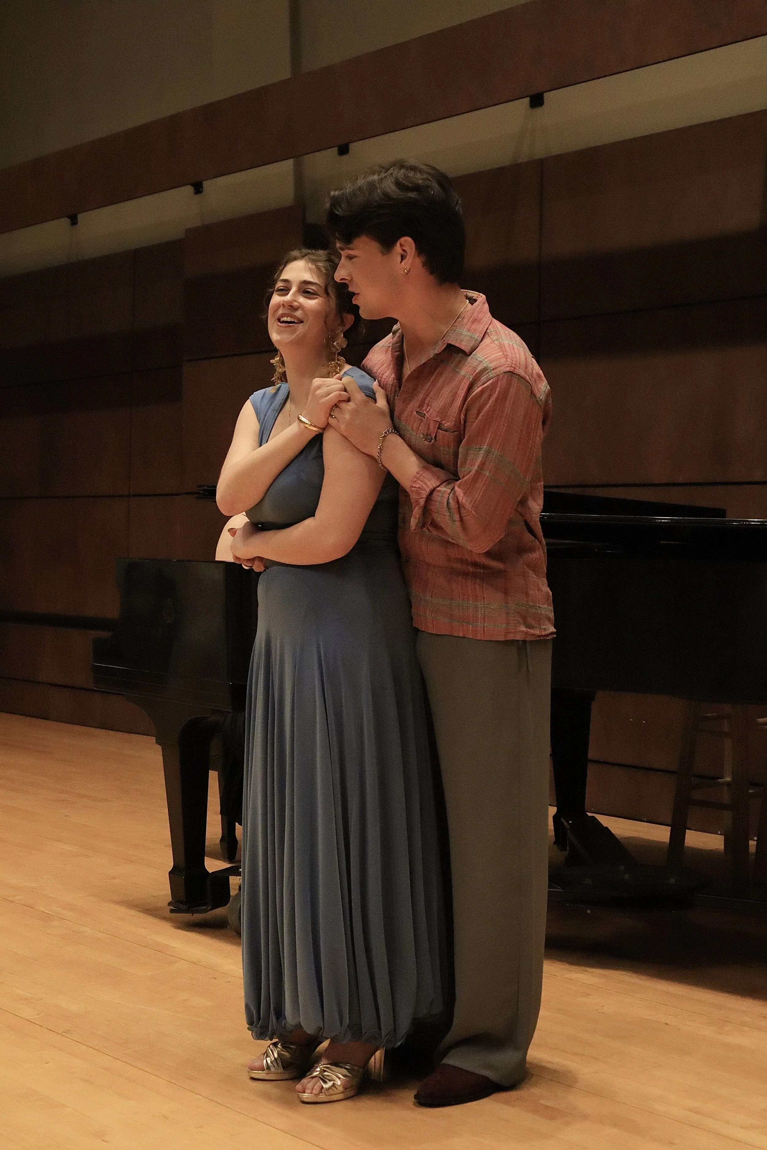 A woman in a long grey dress and gold heels, and a man in a plaid shirt and grey pants, are standing close together, smiling and holding hands in a room with a wood floor, wood paneled walls, and a black grand piano in the background.