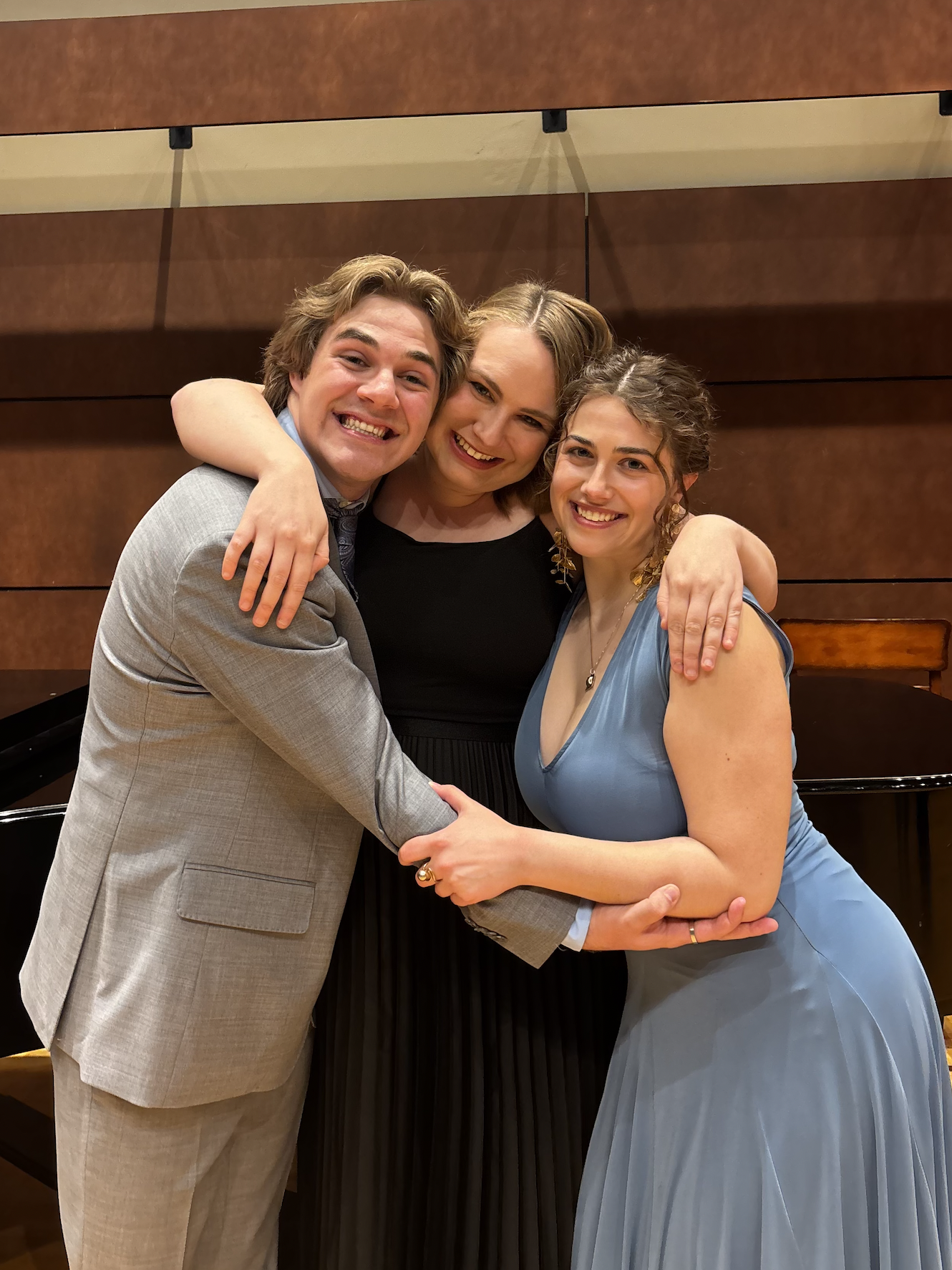Three young adults dressed in formal attire embracing each other and smiling at the camera in front of a wooden wall.