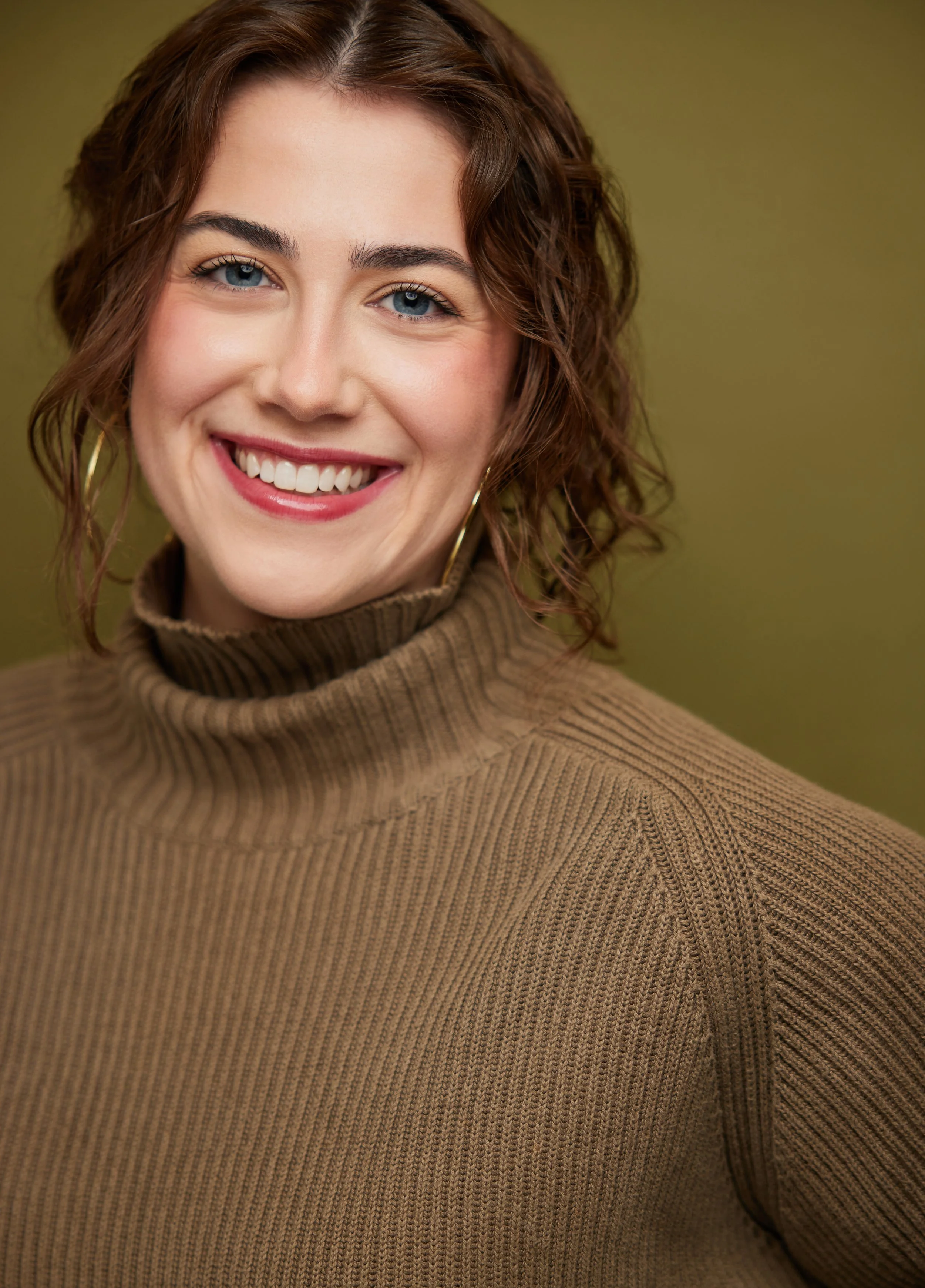 Close-up of a woman with curly brown hair, blue eyes, wearing a brown turtleneck sweater and gold hoop earrings, smiling against a green background.