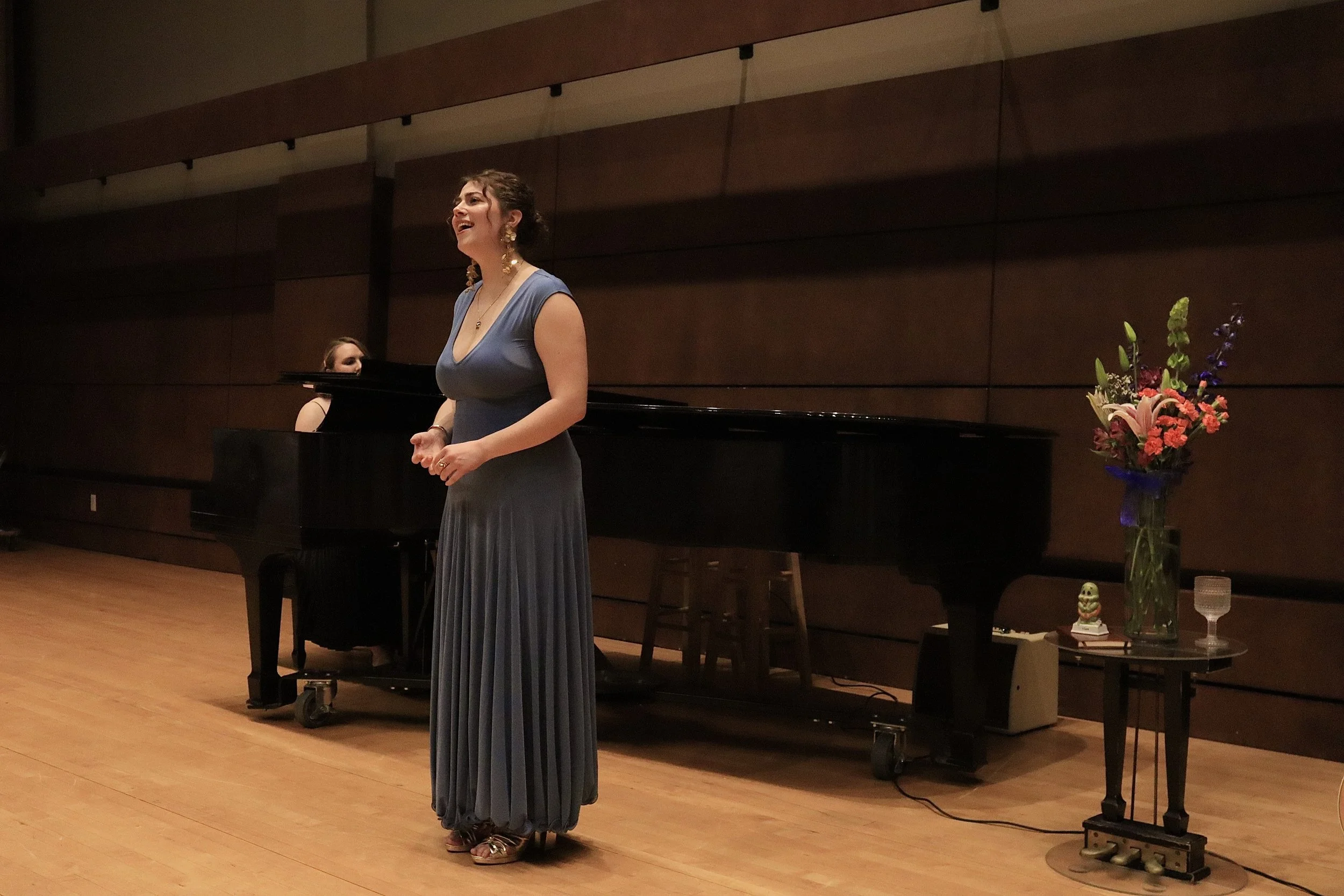 A woman in a blue dress singing on a stage, with a pianist in the background, a flower arrangement on a side table, and wooden paneling on the wall.
