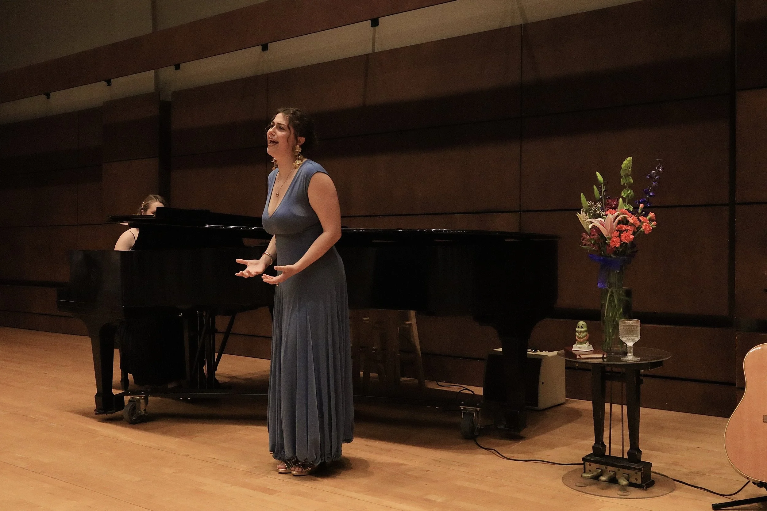 A woman in a long blue dress is singing in a concert hall. Behind her, there is a pianist playing a black grand piano. To her right, there is a table with a vase of flowers, a small decorative figurine, and a glass of water.