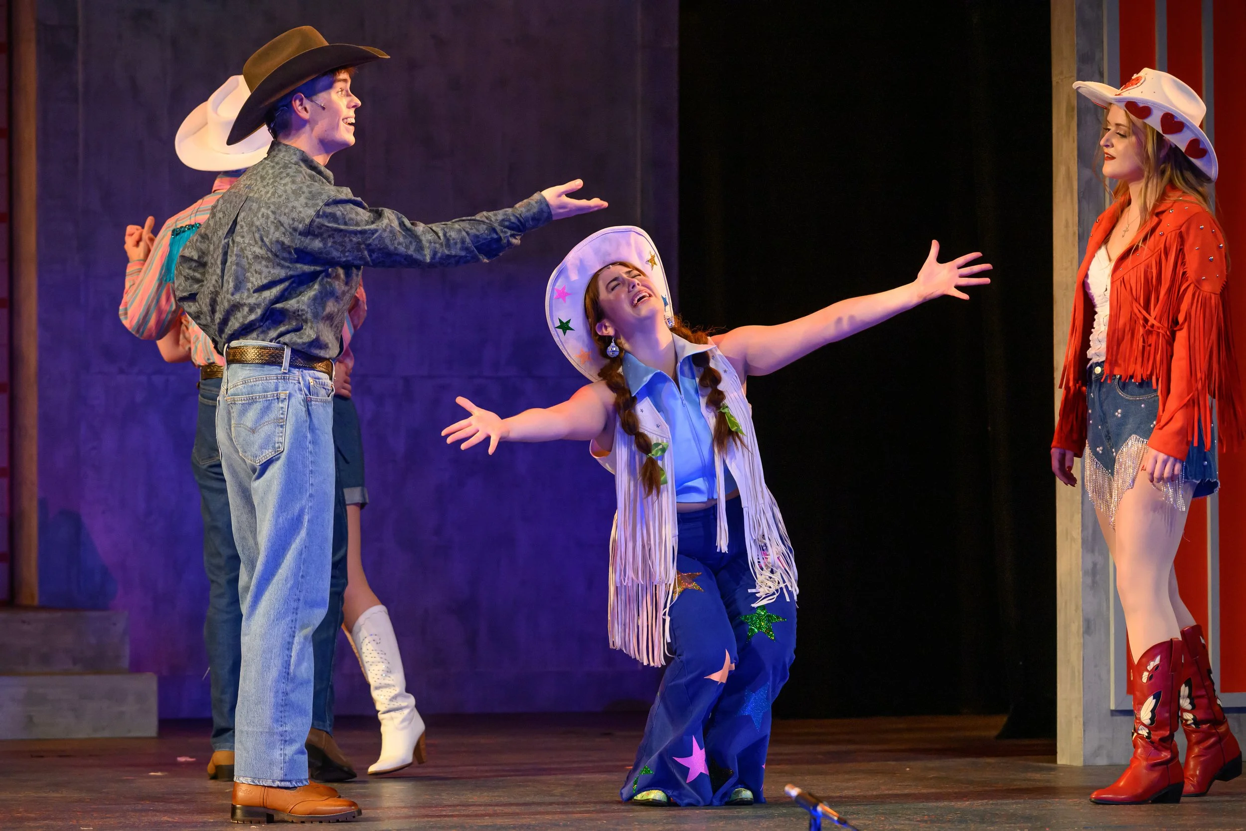 Four women on stage dressed in cowboy and cowgirl costumes, with one woman in the center wearing a star-studded cowboy hat and gesturing toward another woman in a cowboy hat and fringed jacket. They appear to be in the middle of a theatrical performa