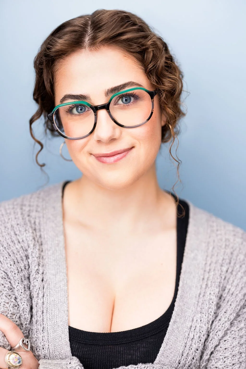 A young woman with curly brown hair, wearing large round glasses with blue and black frames, smiling softly, against a light blue background.