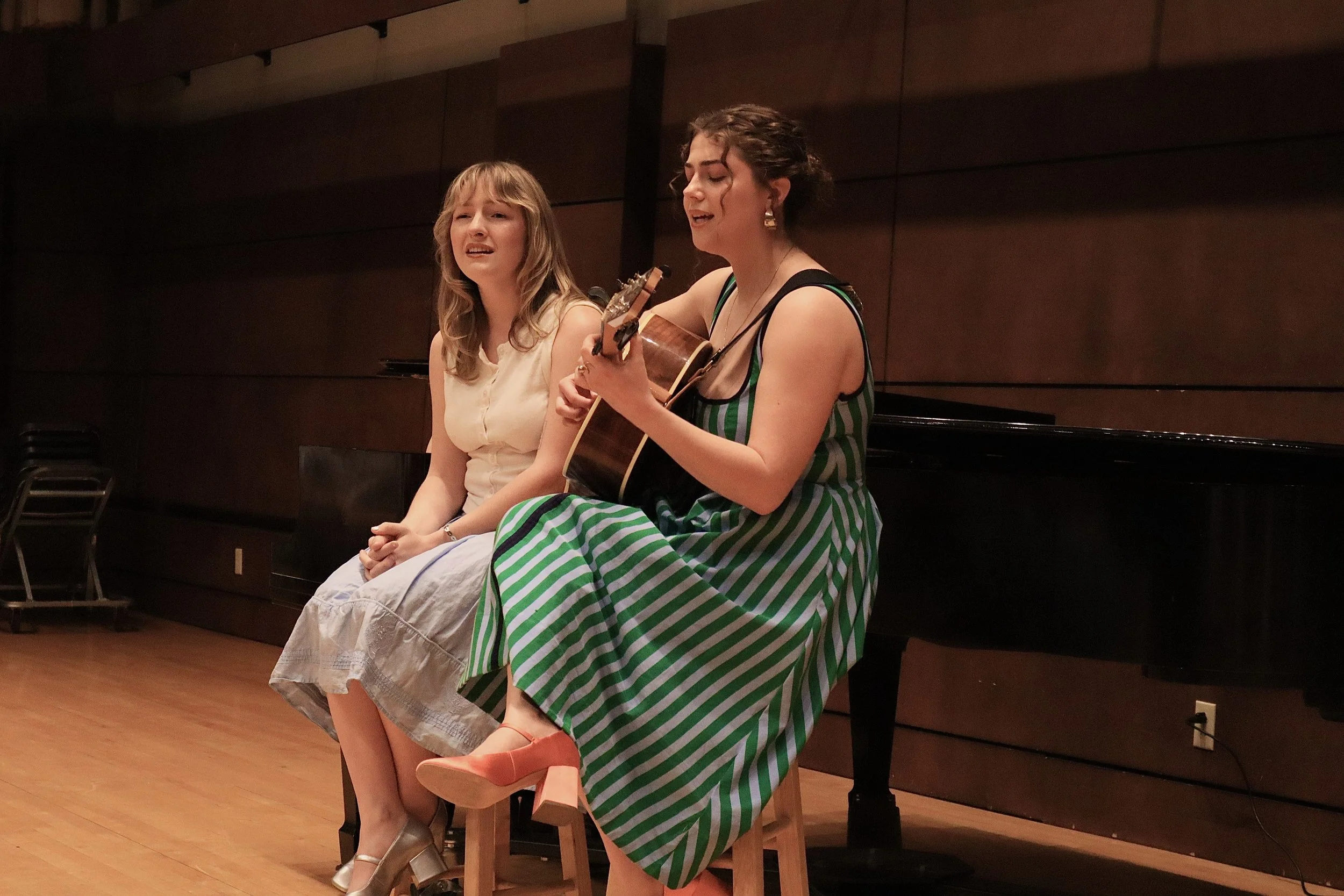Two young women are sitting on stools, with one playing a guitar and singing, in a room with dark wooden walls and a polished wooden floor.