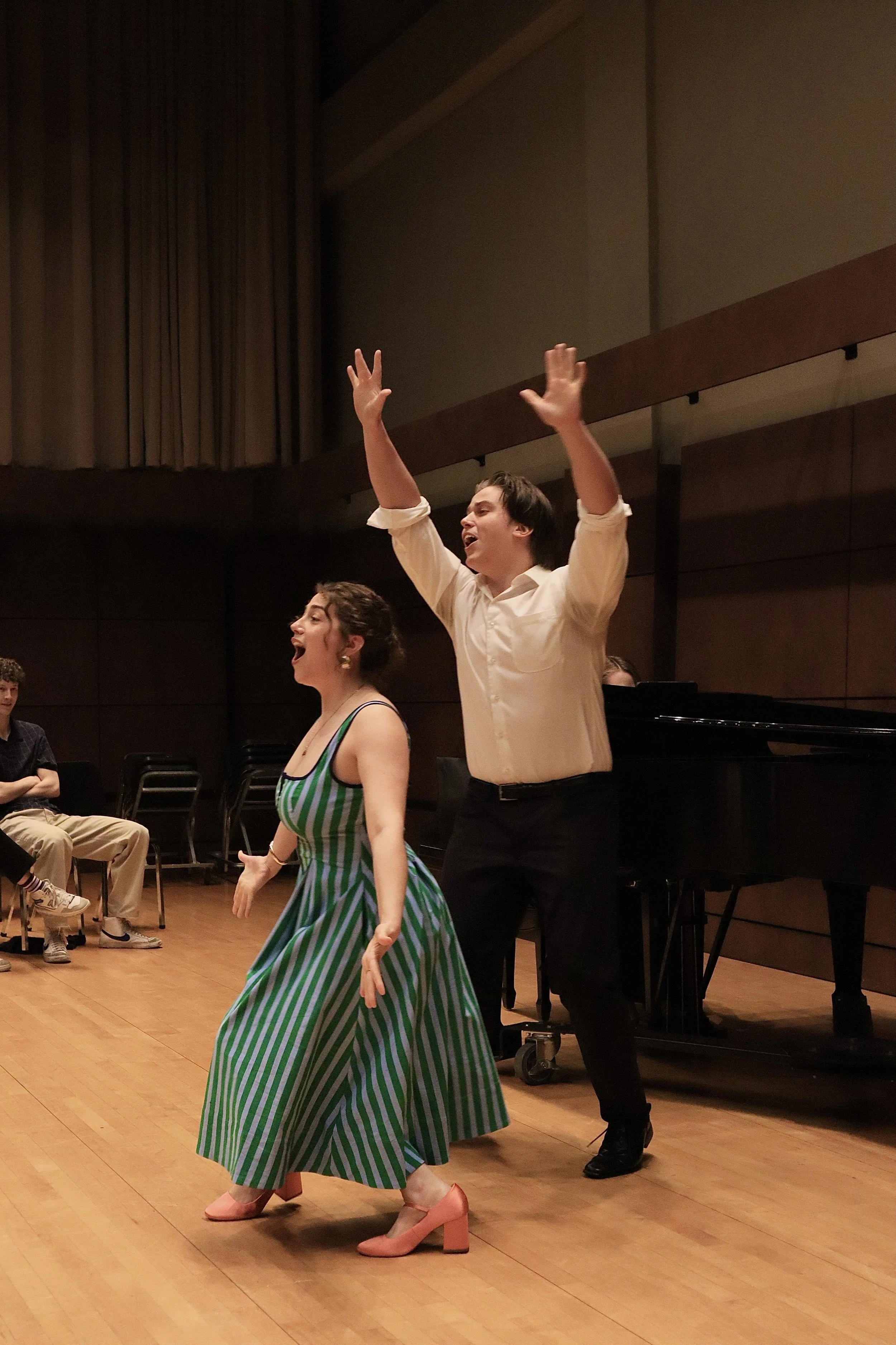 Two performers, a woman in a green and white striped dress and a man in black pants and a white shirt, are dancing and singing on a stage in a concert hall. The woman is wearing pink heels, and the man has his arms raised with a joyful expression. Ot