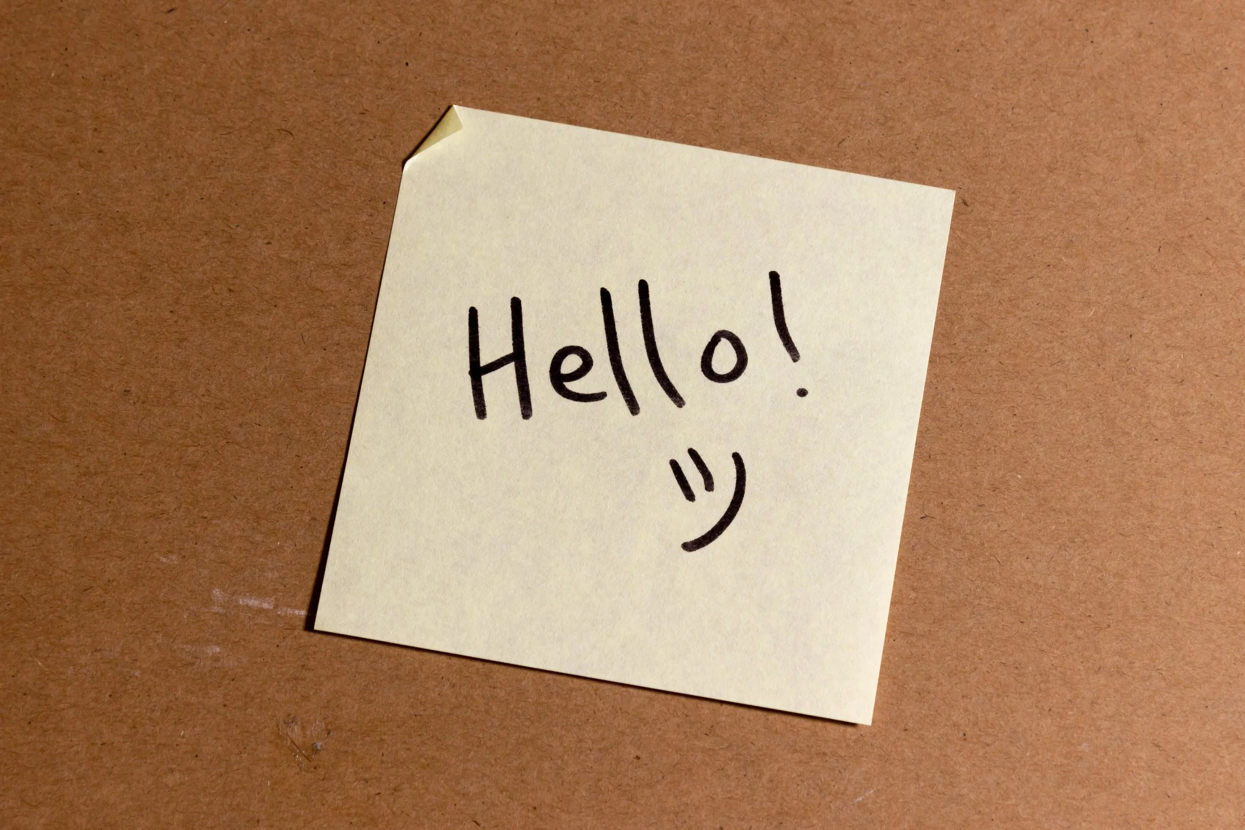 A sticky note with the words 'Hello!' written in black marker and a smiley face, placed on a brown surface.