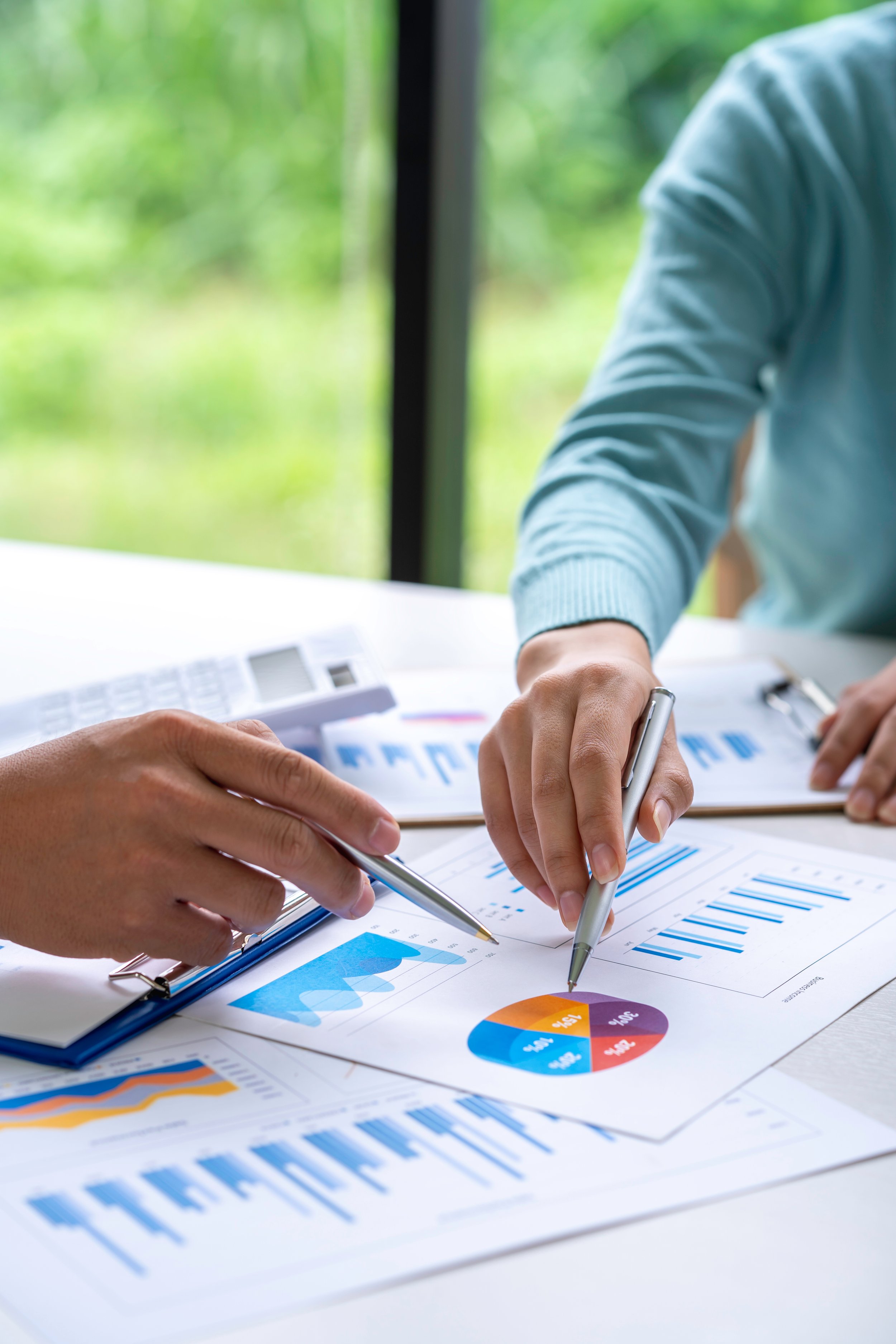 Two people analyzing business graphs and pie charts on papers and a clipboard at a desk with a calculator, with greenery visible through a window in the background.