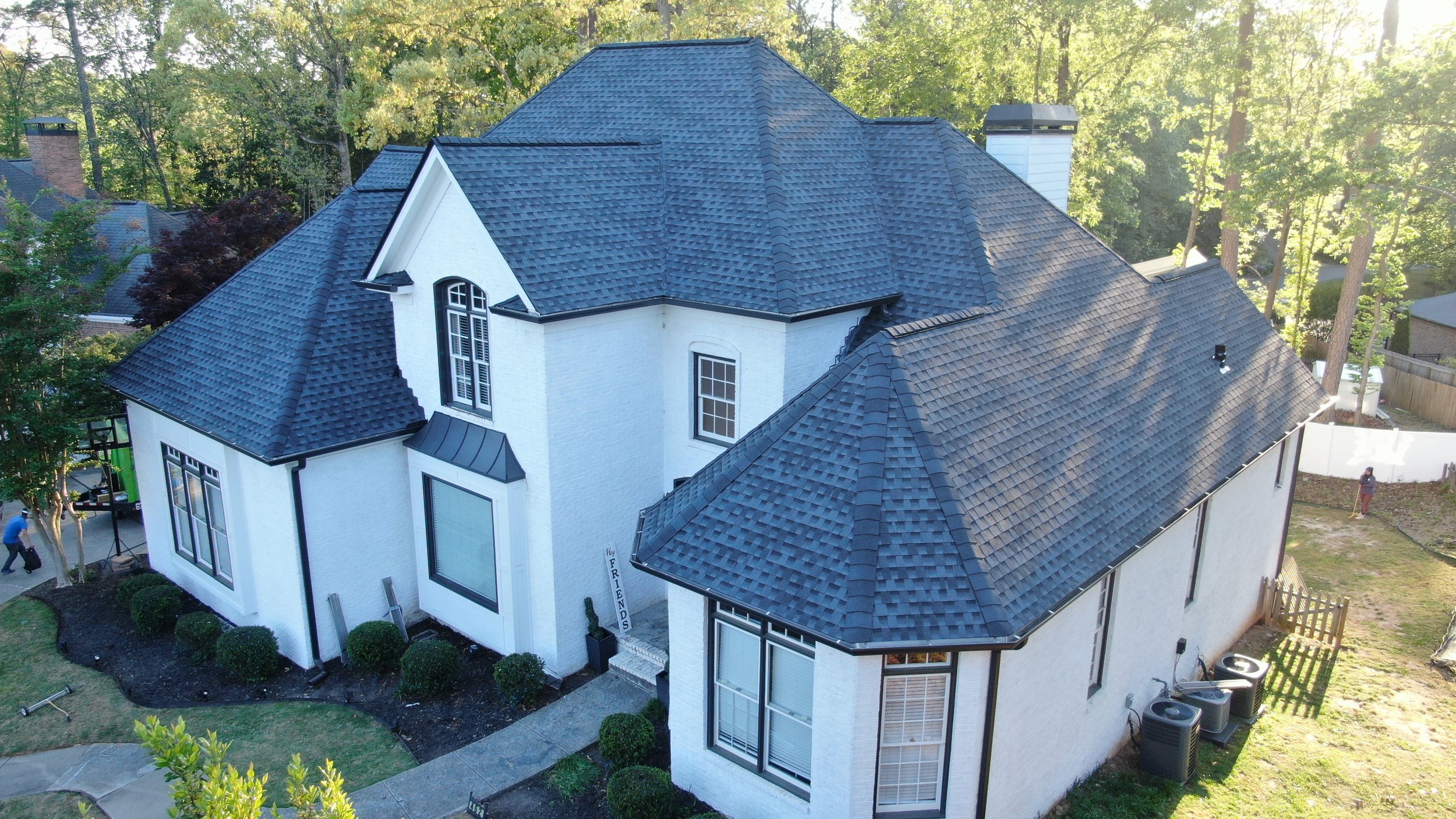 A large white house with black trim and a dark shingled roof, surrounded by trees, with a backyard, lawn, and people working.