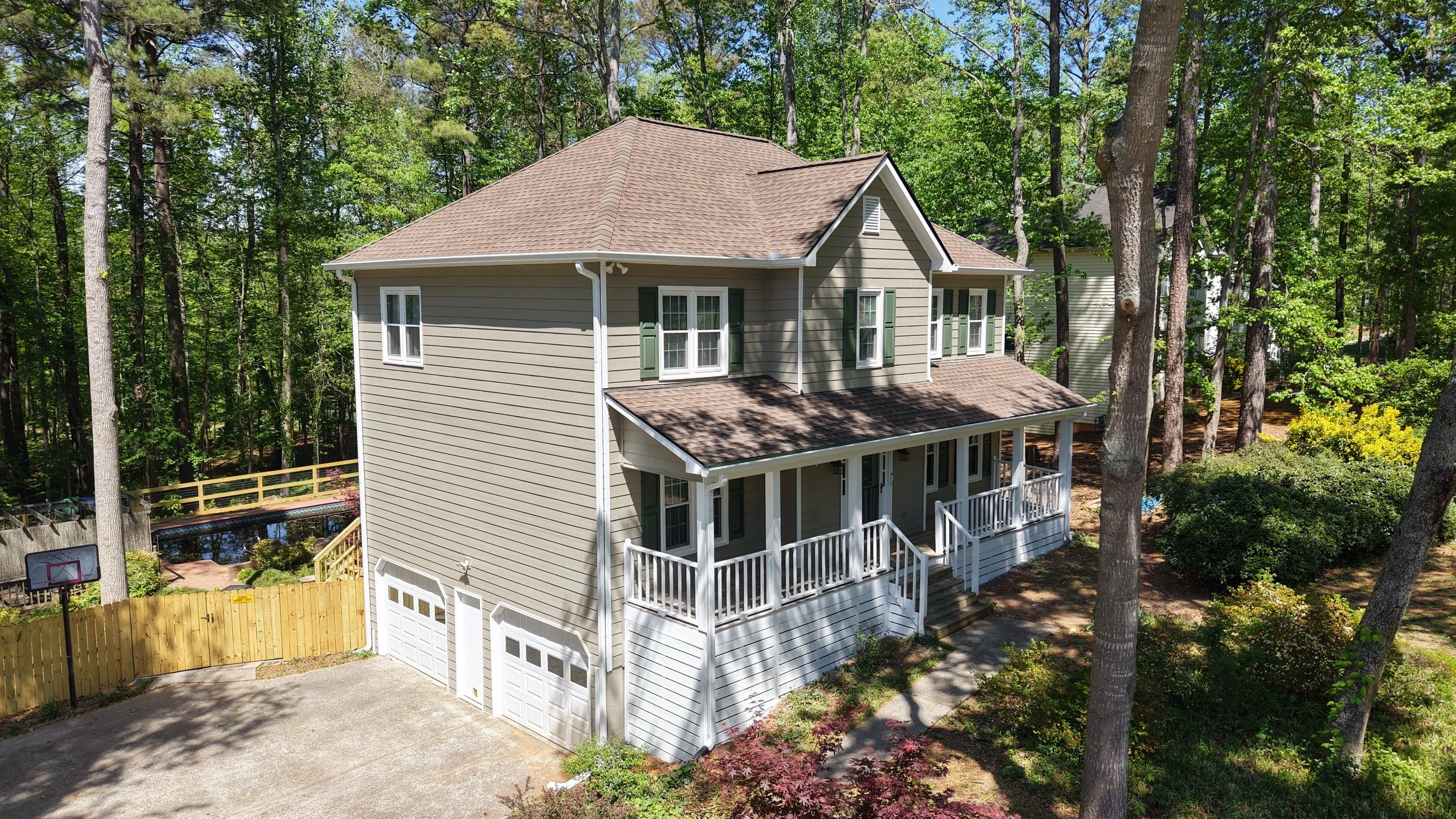 A two-story house with beige siding, green shutters, and a brown shingled roof, surrounded by tall trees in a wooded area, with a porch, driveway, and fenced yard.