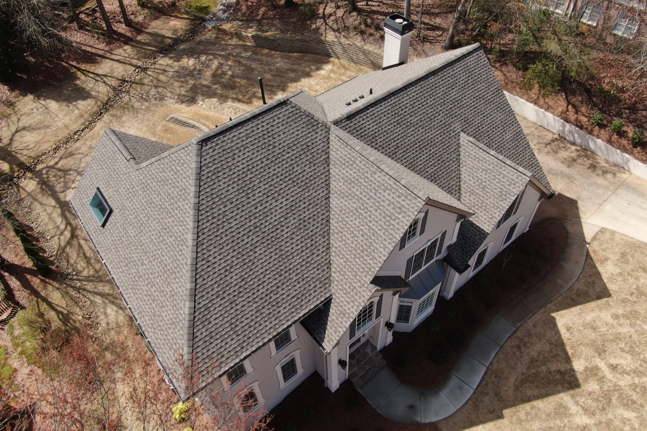Aerial view of a house with a grey tiled roof, multiple gable peaks, and a chimney, surrounded by trees and a backyard with a concrete patio and landscaped areas.