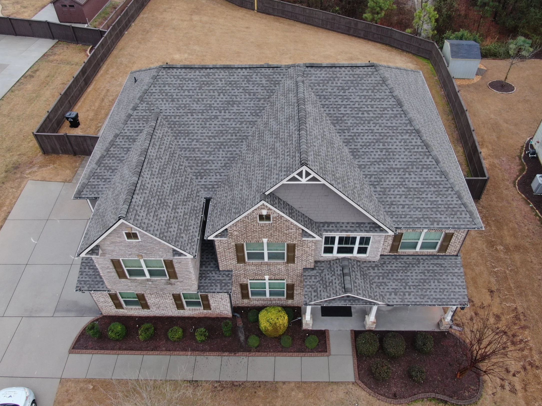 Aerial view of a house with a gray shingled roof, brick exterior walls, and multiple windows, surrounded by a fenced backyard with brown grass, small shrubs, and a concrete driveway.
