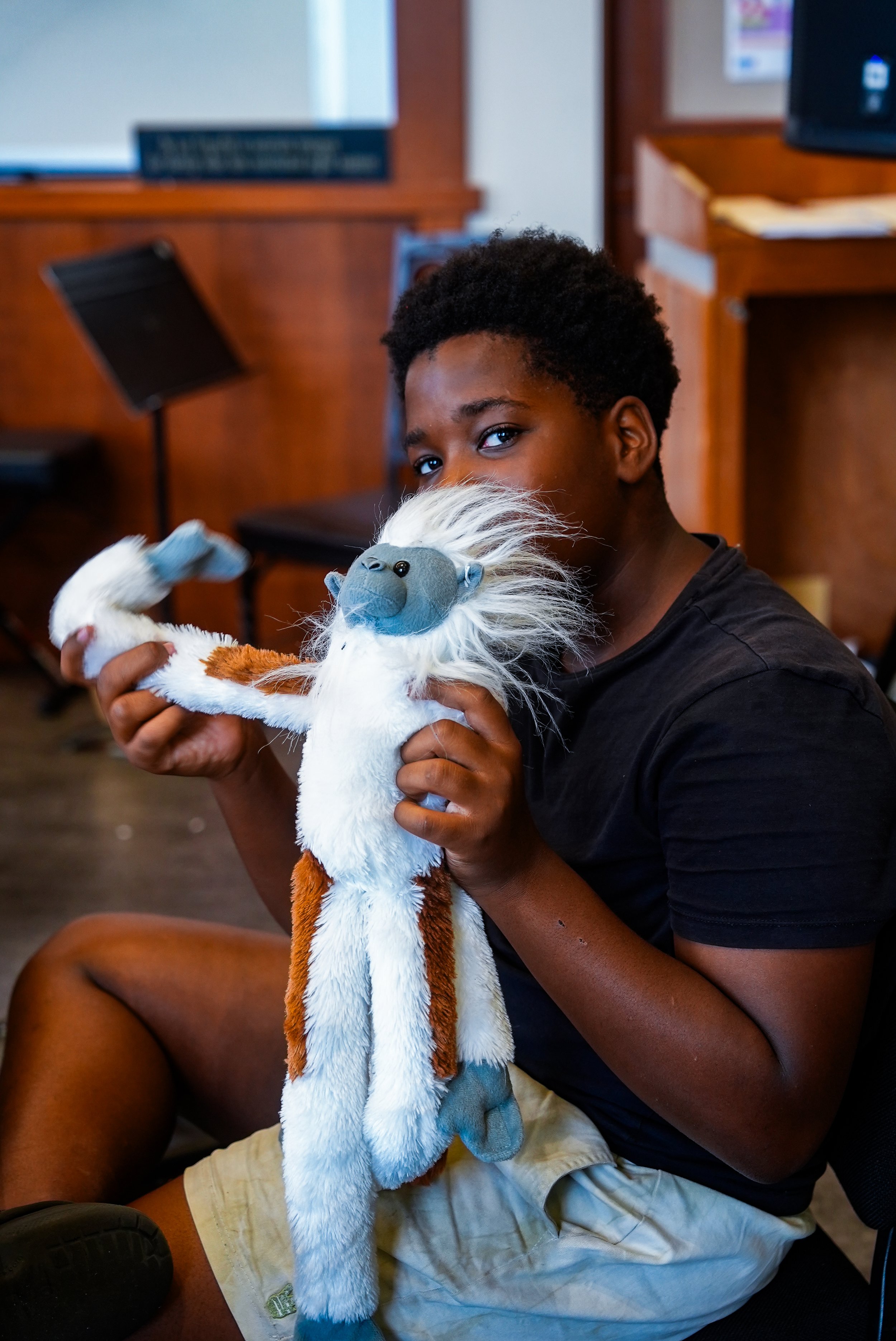 A boy with short curly hair sitting in a room with wooden furniture, holding and cuddling a plush monkey toy with a light blue face, white mane, and brown and white body.