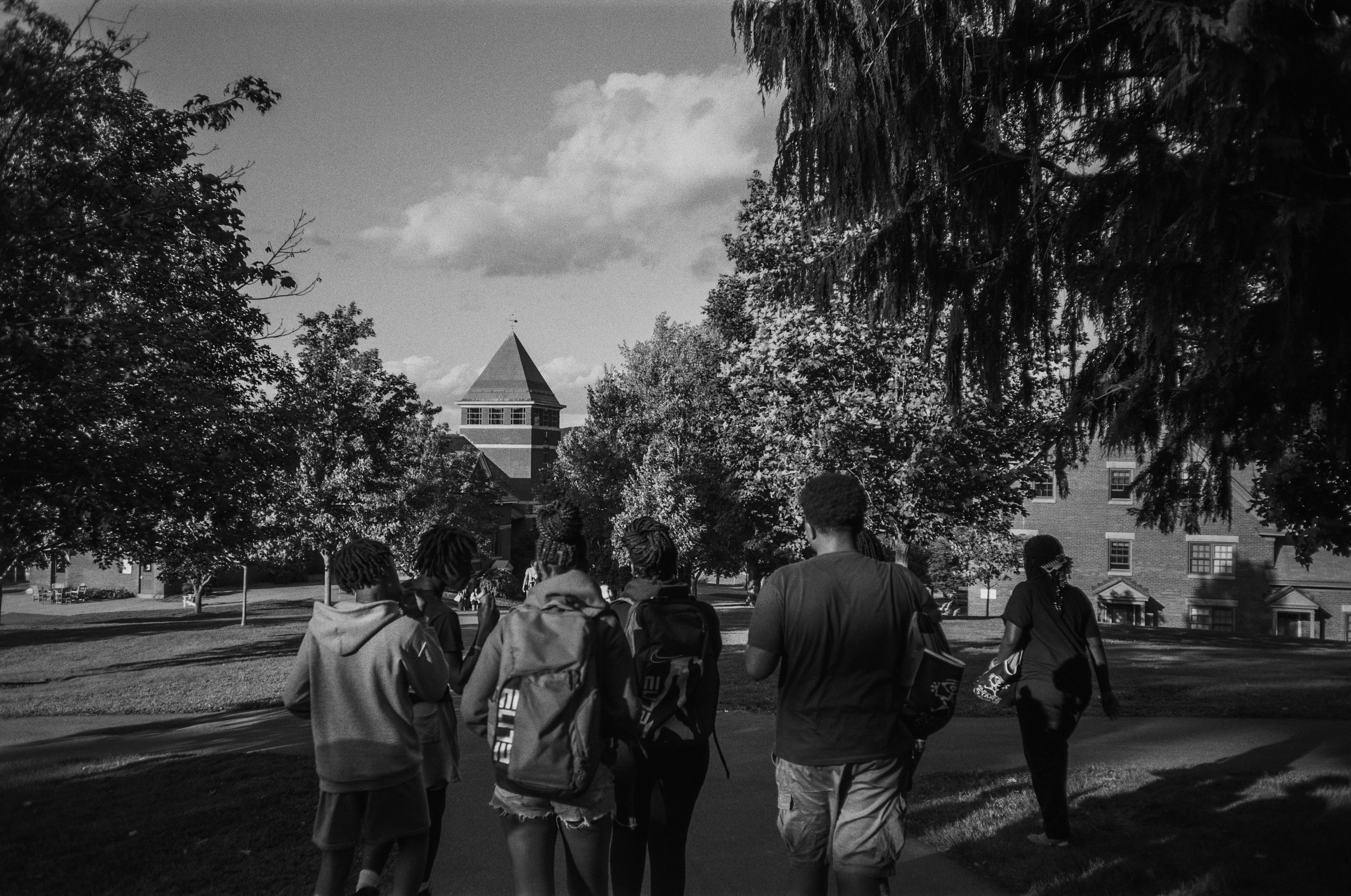 Group of young people walking outside on a college campus, surrounded by trees and buildings, with a tower in the background.