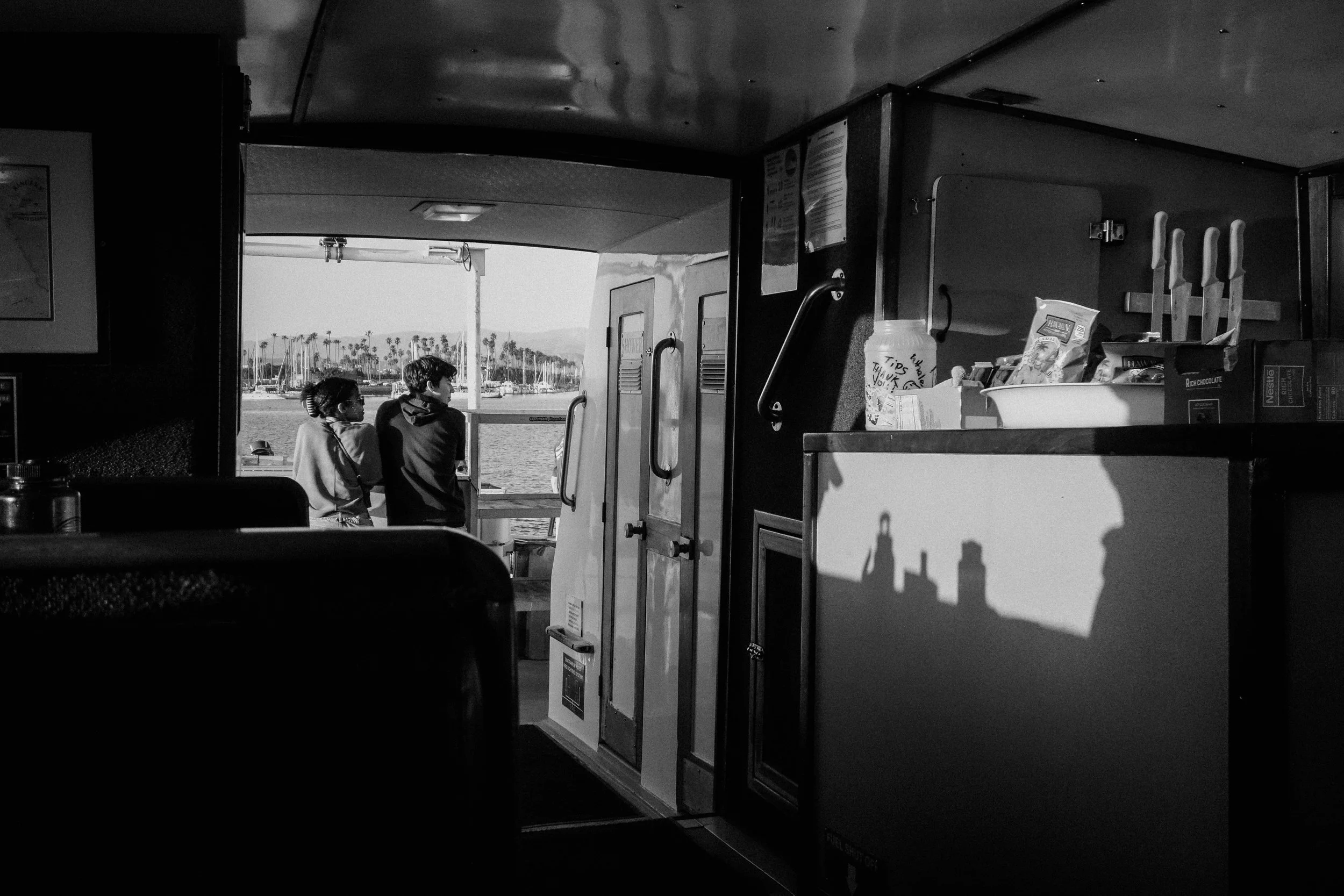 Two children stand on a boat deck, gazing at the water and shoreline with palm trees in the distance, seen through an open window from inside the boat.