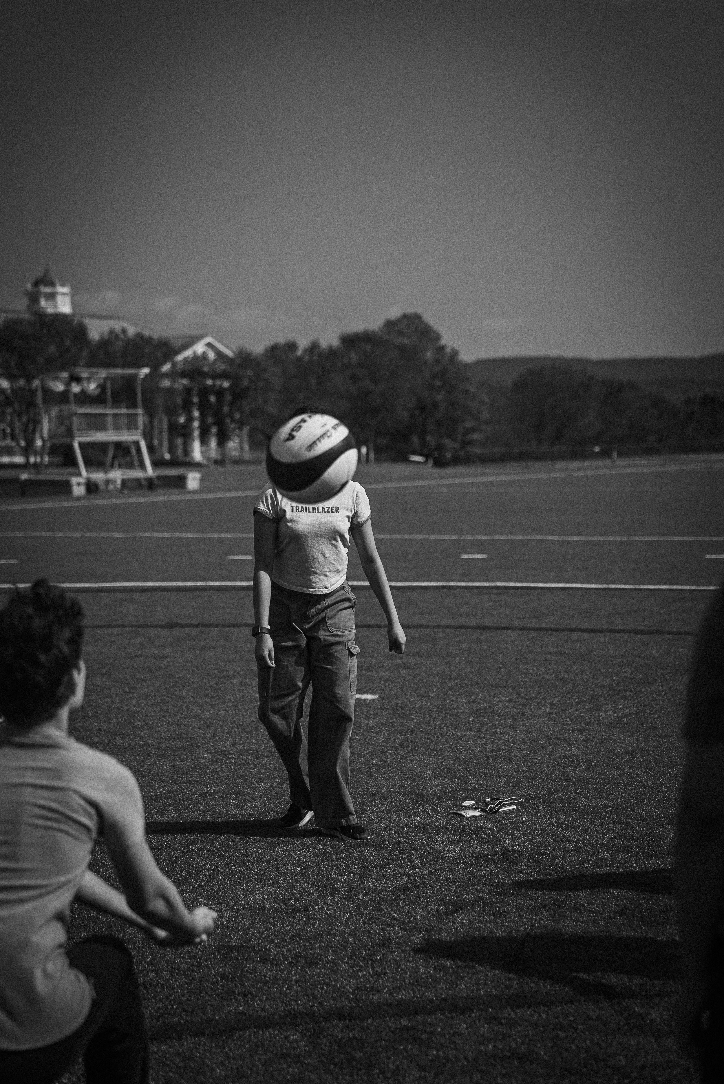 A child with a helmet on their head standing on a sports field, with other children sitting nearby, during daytime.