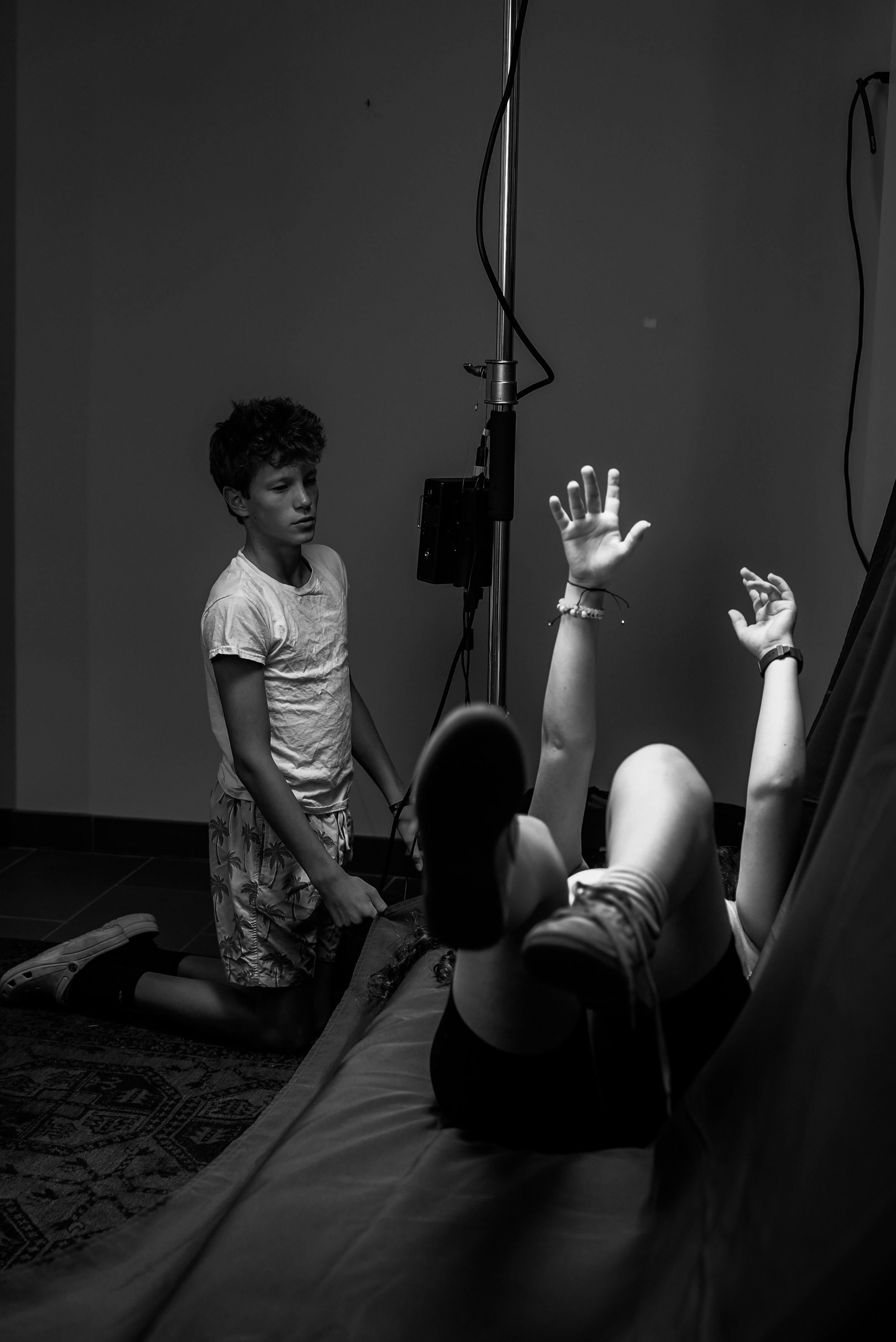 A young boy kneeling next to a girl lying on a bed, with her legs bent and arms raised. The girl appears to be reaching out or gesturing. The scene takes place in a dimly lit room with a plain wall and some equipment hanging from a pole.