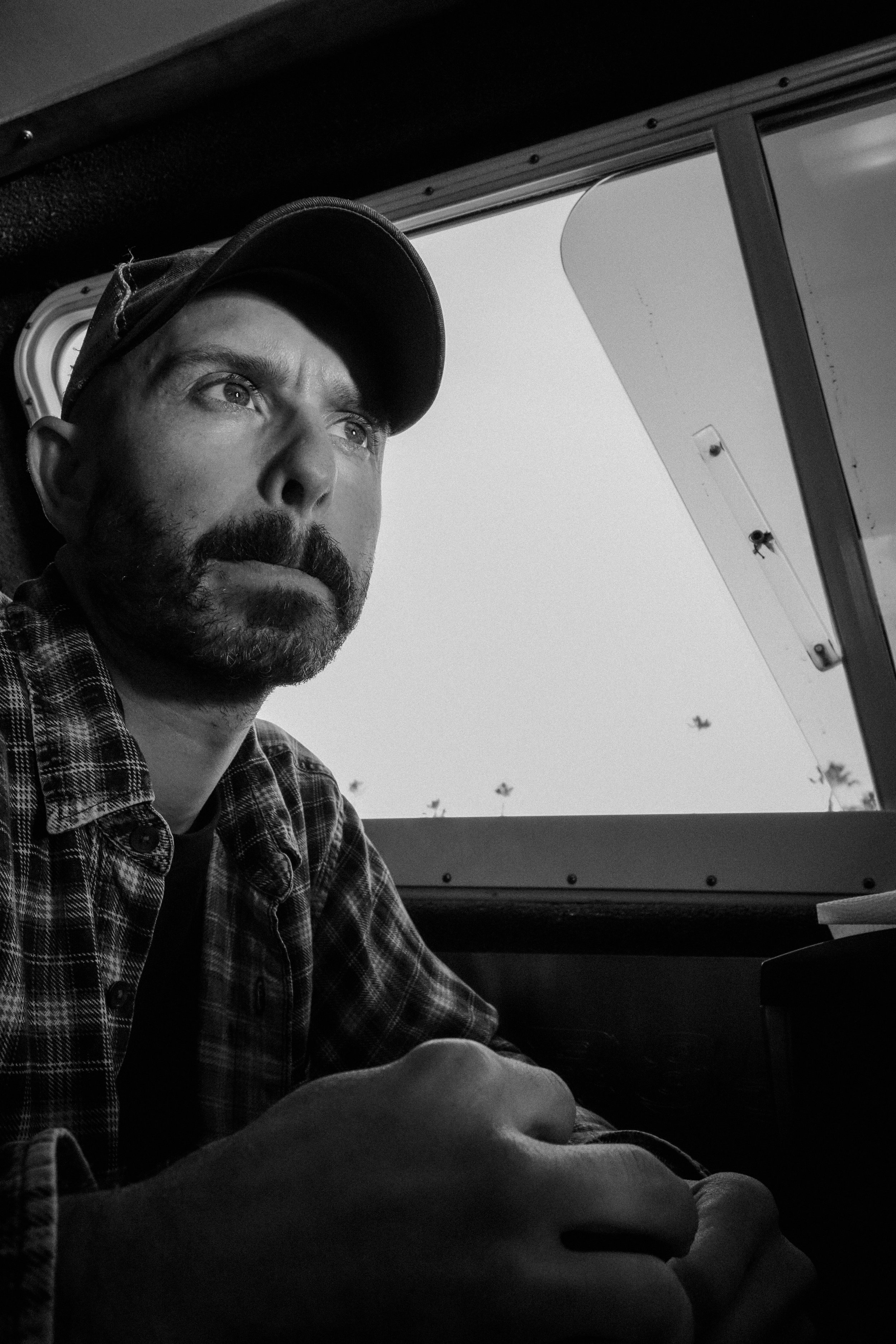 A man with a beard wearing a cap and plaid shirt sitting inside a vehicle, looking thoughtfully out the window at a clear sky with a few birds flying.