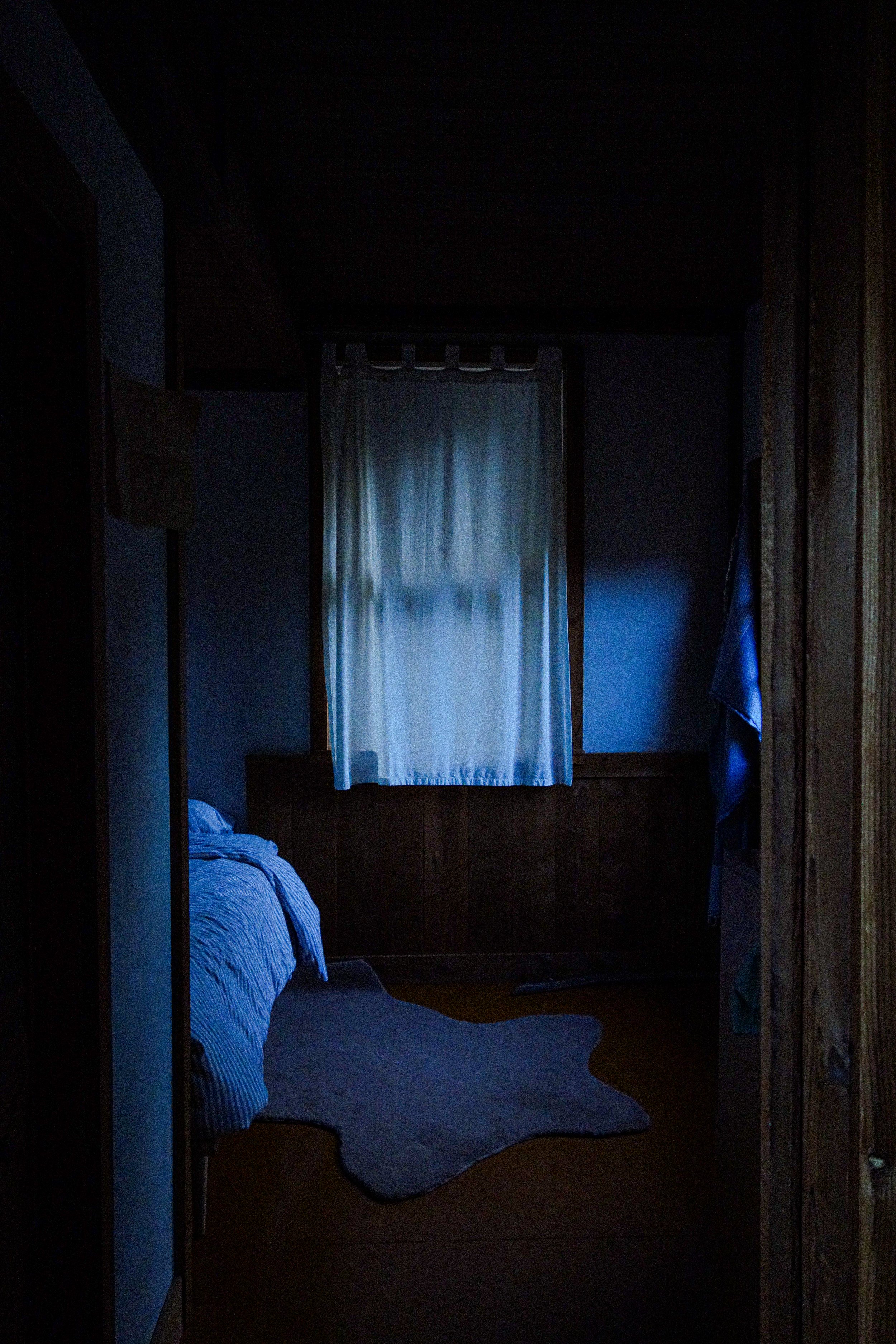 A dimly lit bedroom with a bed, a window with closed curtains, and a small rug on the floor.