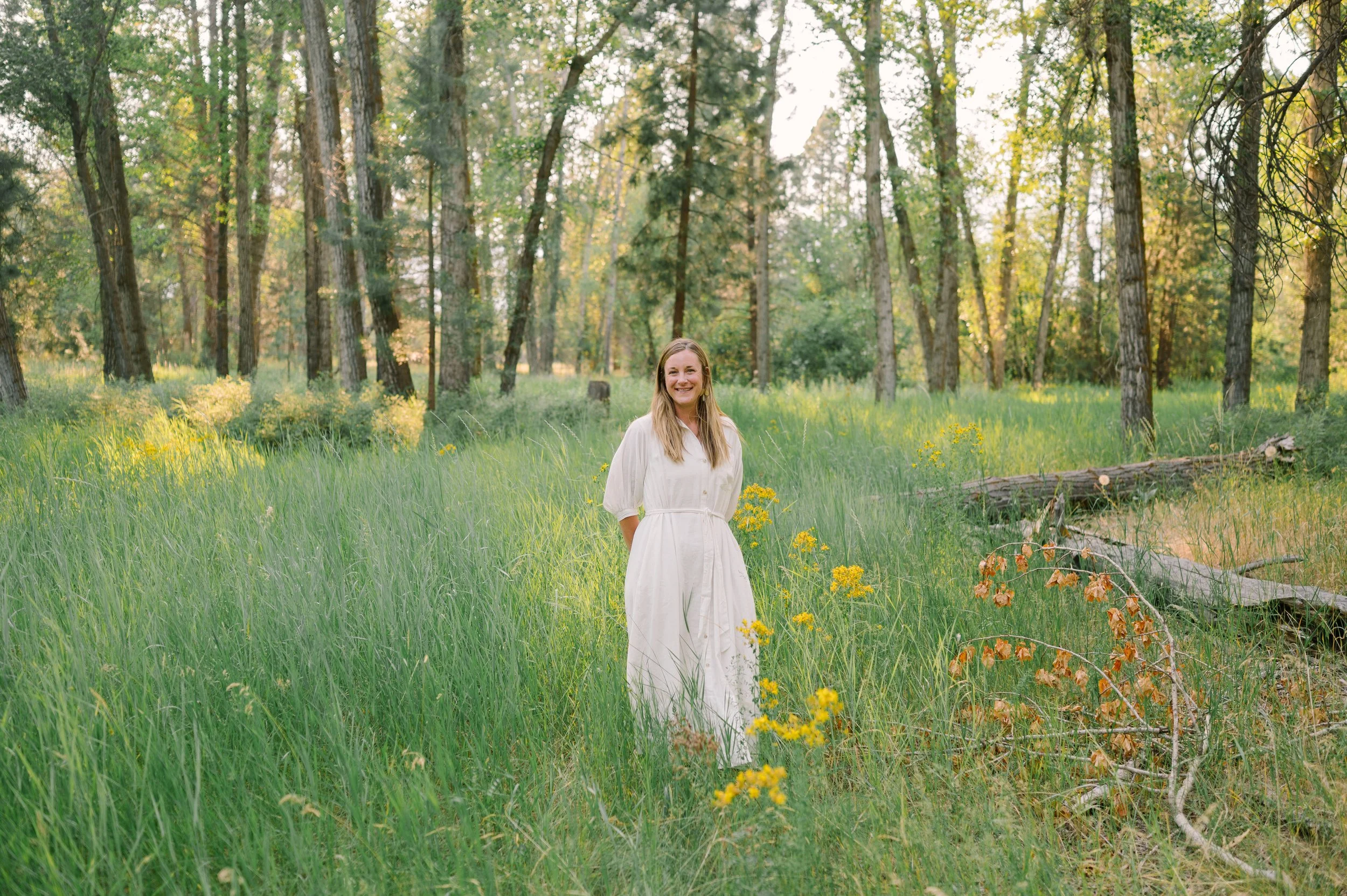 A woman in a white dress smiling and standing in a lush green forest with tall trees and sunlight filtering through the leaves.