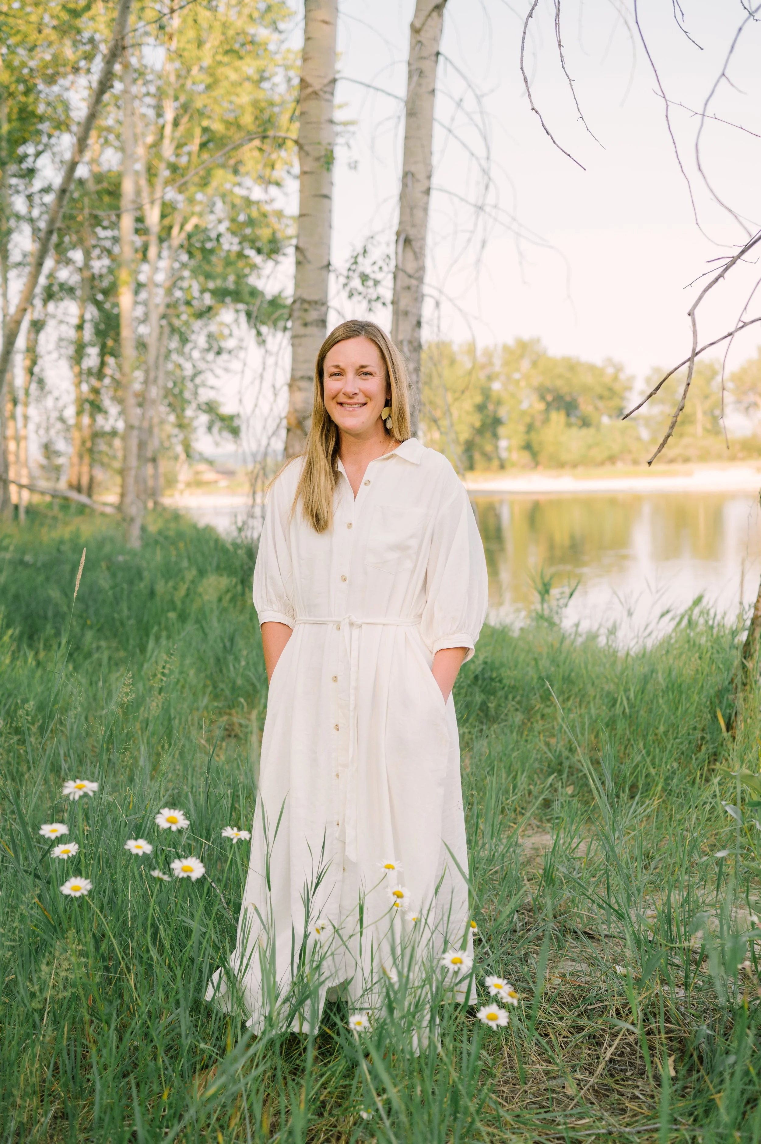 A woman smiling outdoors in a white dress surrounded by green grass and wildflowers, near a body of water with trees in the background.