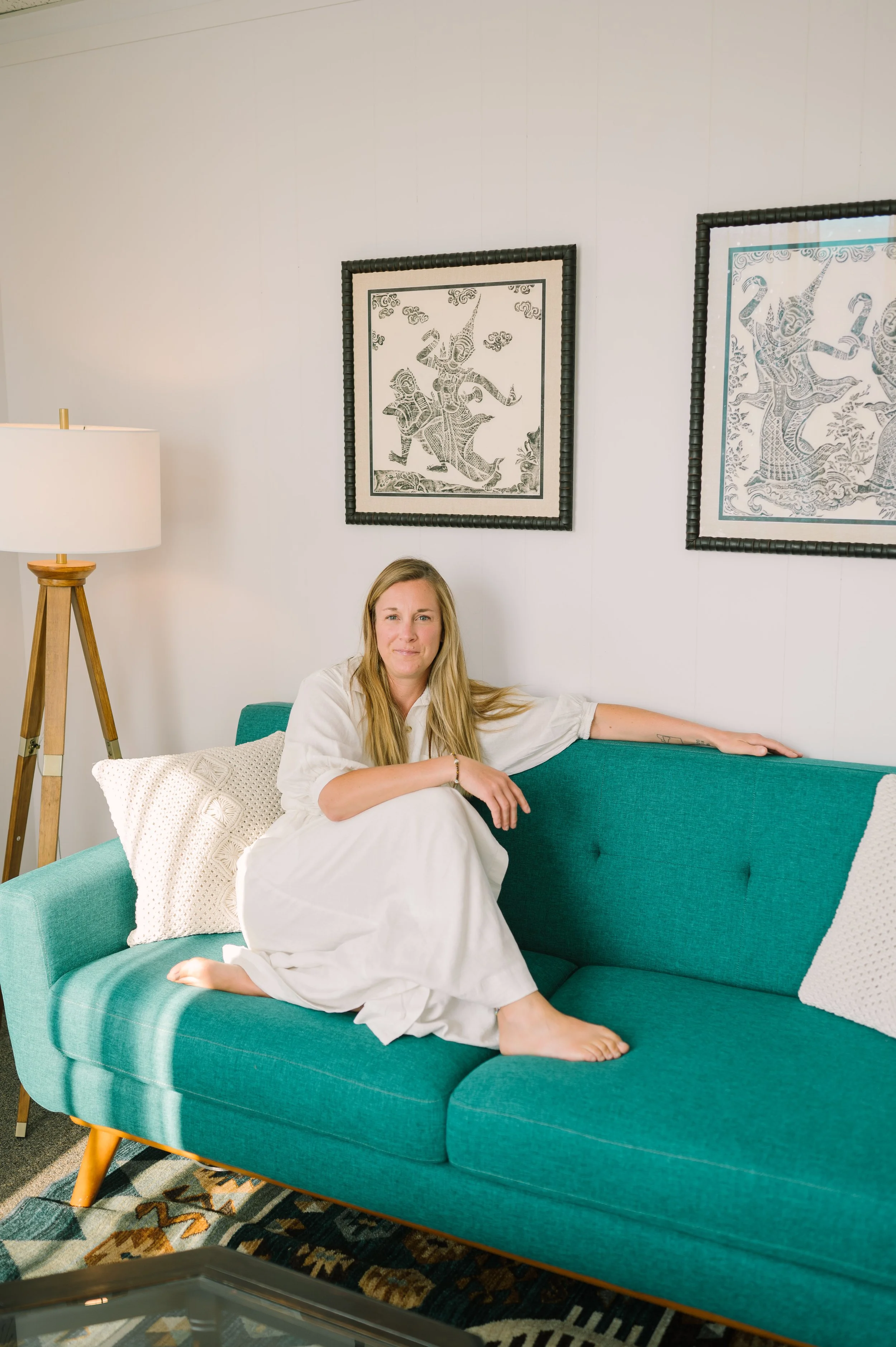 A woman with long blonde hair sitting on a teal couch in a living room. She is wearing a white dress and is barefoot, with one leg tucked under the other. There are two decorative pillows on the couch, one textured and one plain. Behind her, a wall features two framed black-and-white artworks depicting traditional dance figures. To the left, there is a wooden tripod floor lamp with a large white lampshade.