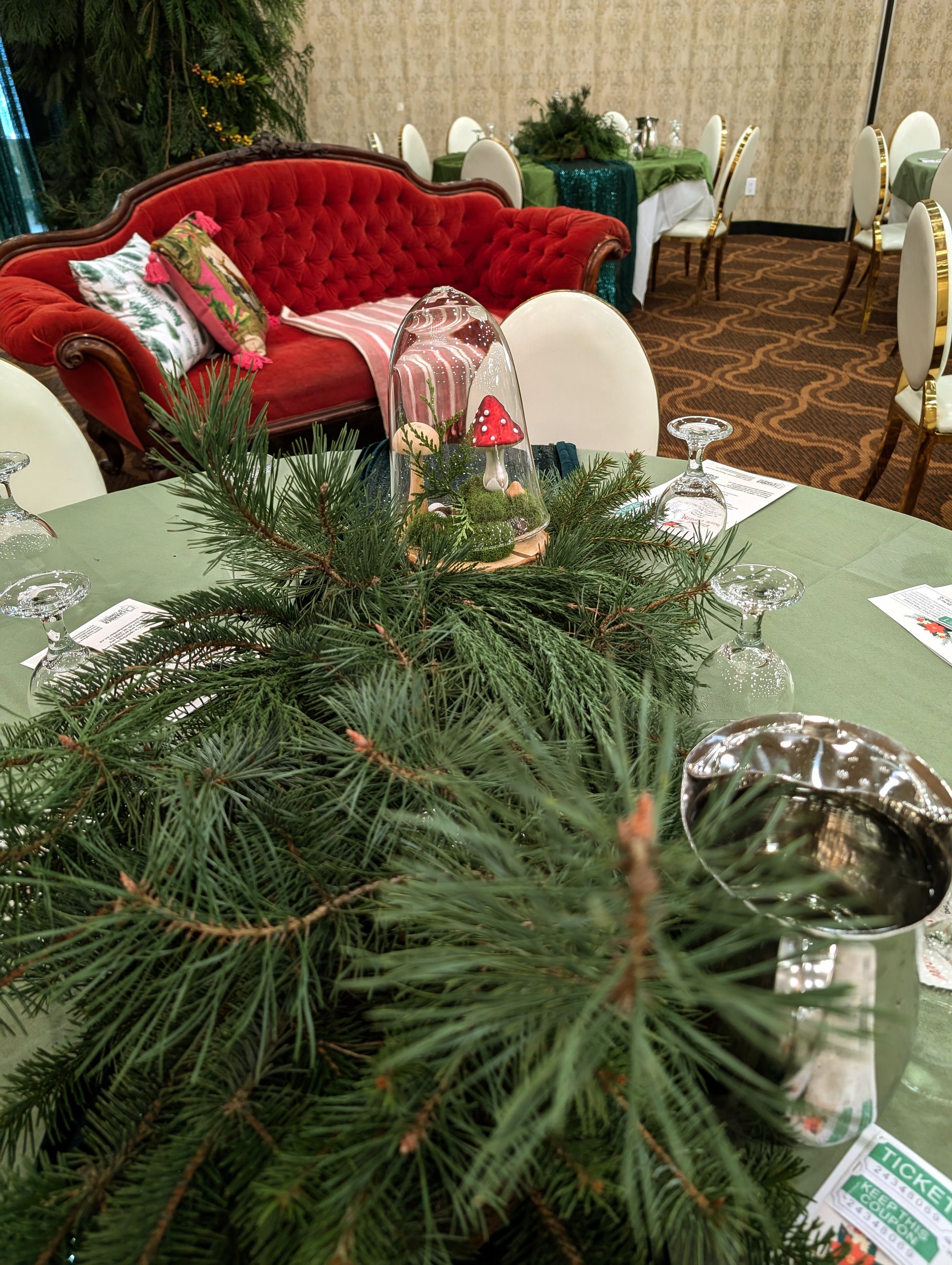 A decorated holiday table with pine branch centerpiece, glass dome with mushroom ornament, and place settings, with a red vintage sofa and additional tables in the background.