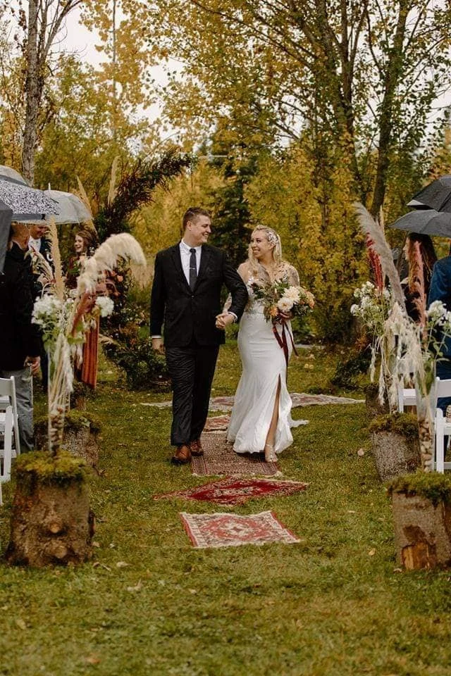 A bride and groom walk down an outdoor aisle replacing an autumn scene with trees displaying fall foliage, surrounded by wedding guests holding umbrellas.
