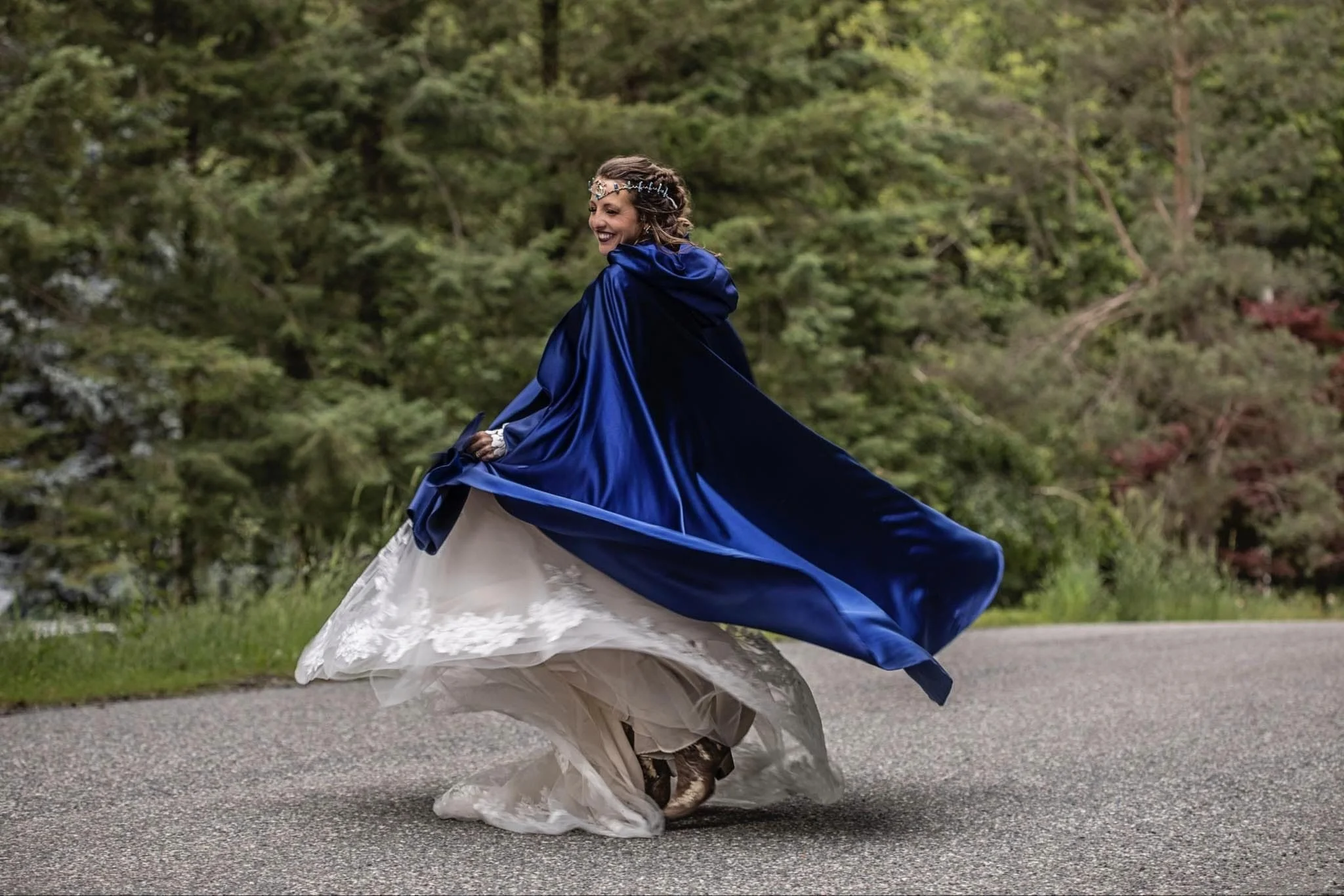 A woman in a wedding dress twirling on a paved path, wearing a blue cape and a floral headband, with a green wooded background.
