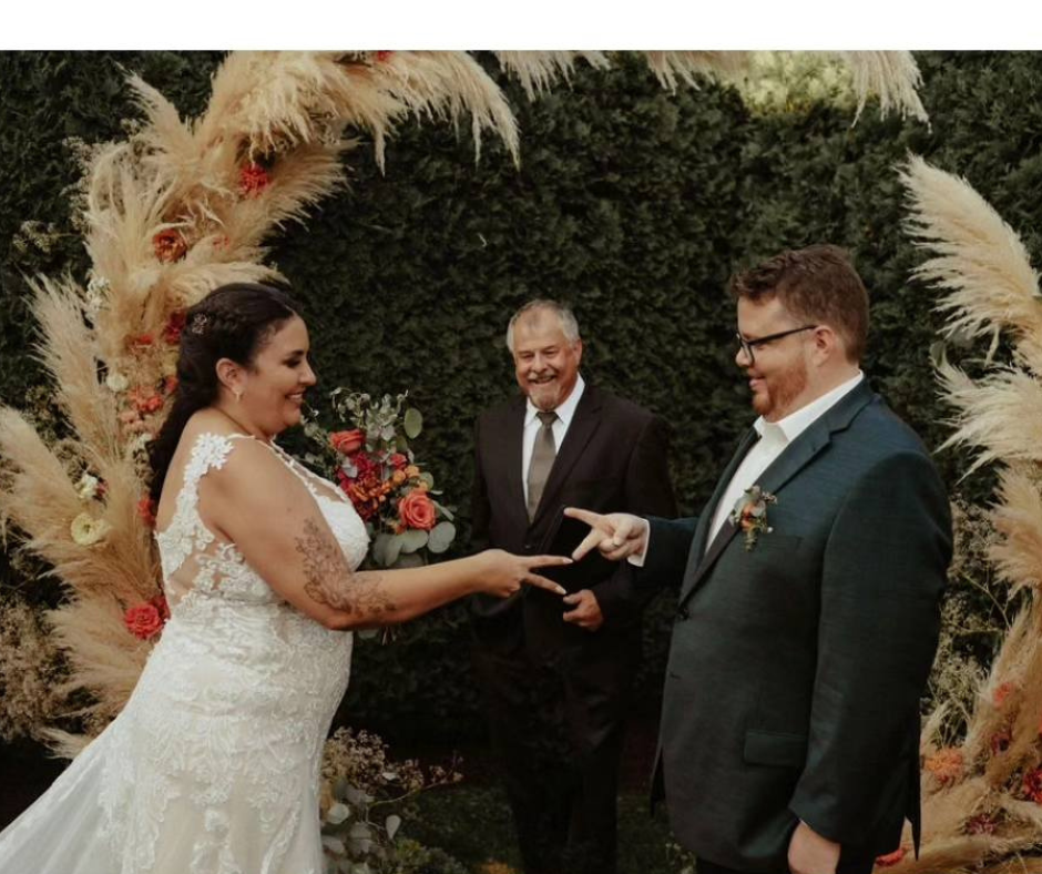 A bride and groom exchange rings during their wedding ceremony outdoors, with an officiant smiling in the background, surrounded by decorative pampas grass and flowers.
