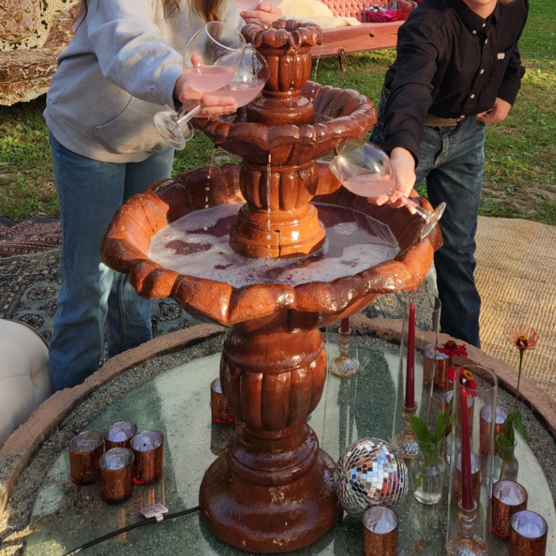 Two people filling wine glasses from a tiered fountain in a garden setting, with decorative items and flowers nearby.