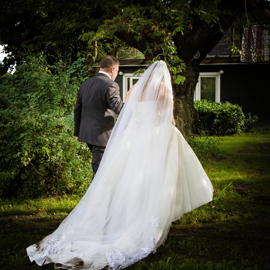 Wedding couple walking outdoors, the bride in a white wedding dress with a veil, the groom in a dark suit, standing near a large tree with greenery and a house in the background.